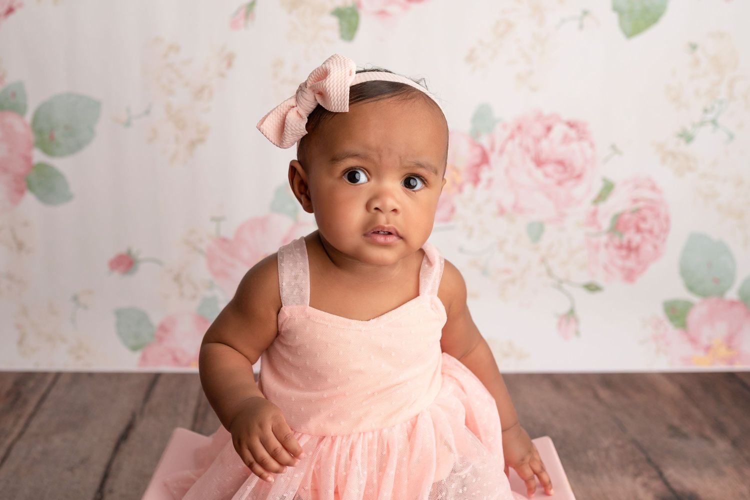 Adorable baby girl in pink tutu dress with bow headband sits against floral backdrop for professional portrait photo.