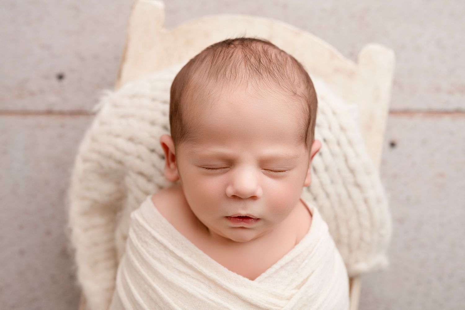 Peaceful sleeping newborn wrapped in soft cream-colored fabric against light wooden backdrop.