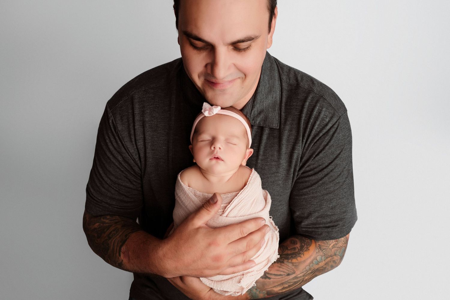 A person in a dark gray t-shirt tenderly holds and kisses a swaddled newborn baby against a light gray background.
