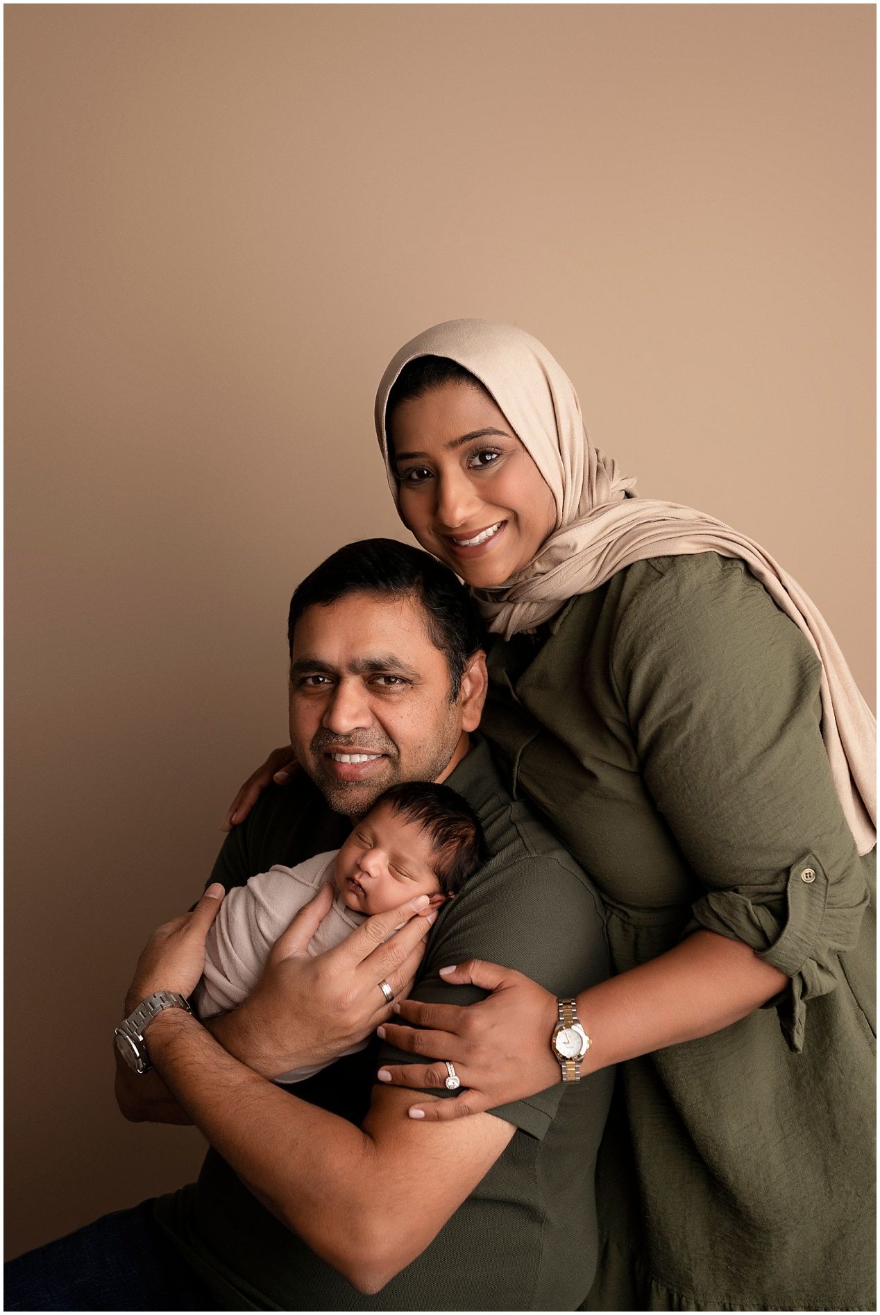 A family portrait against a neutral background shows parents in casual attire holding their newborn baby.