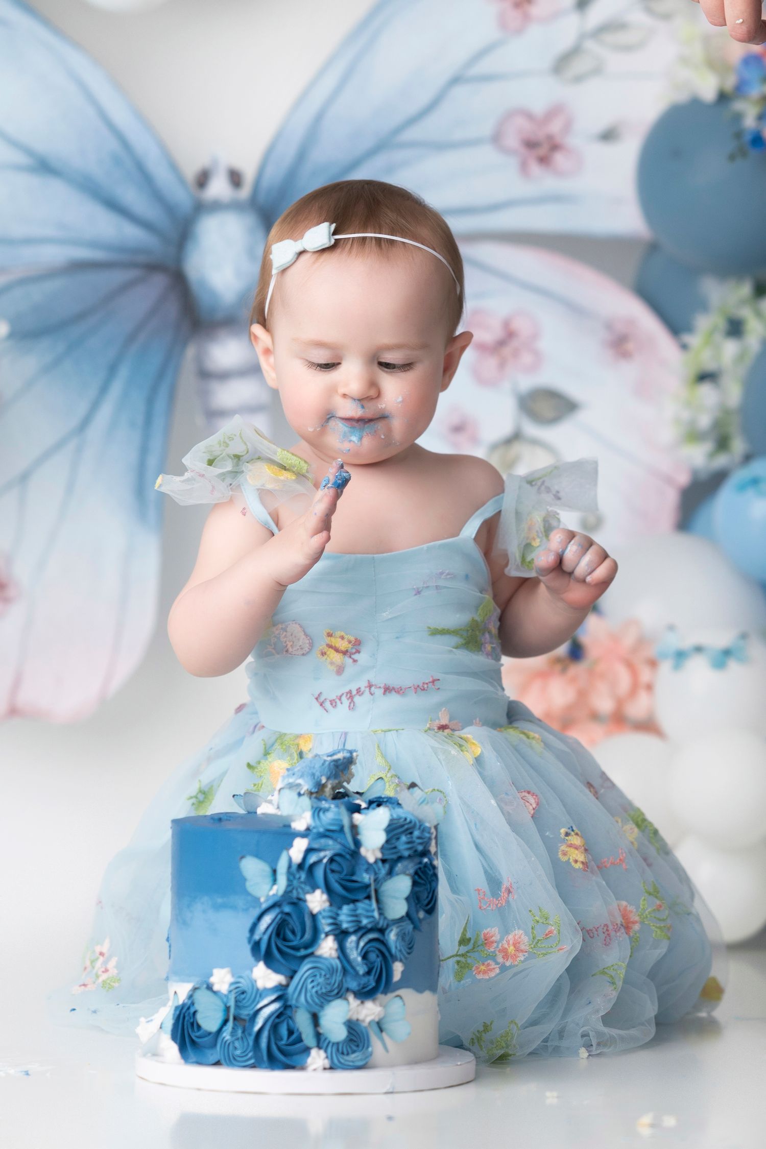 Baby in floral dress sits next to blue smash cake during first birthday photoshoot.