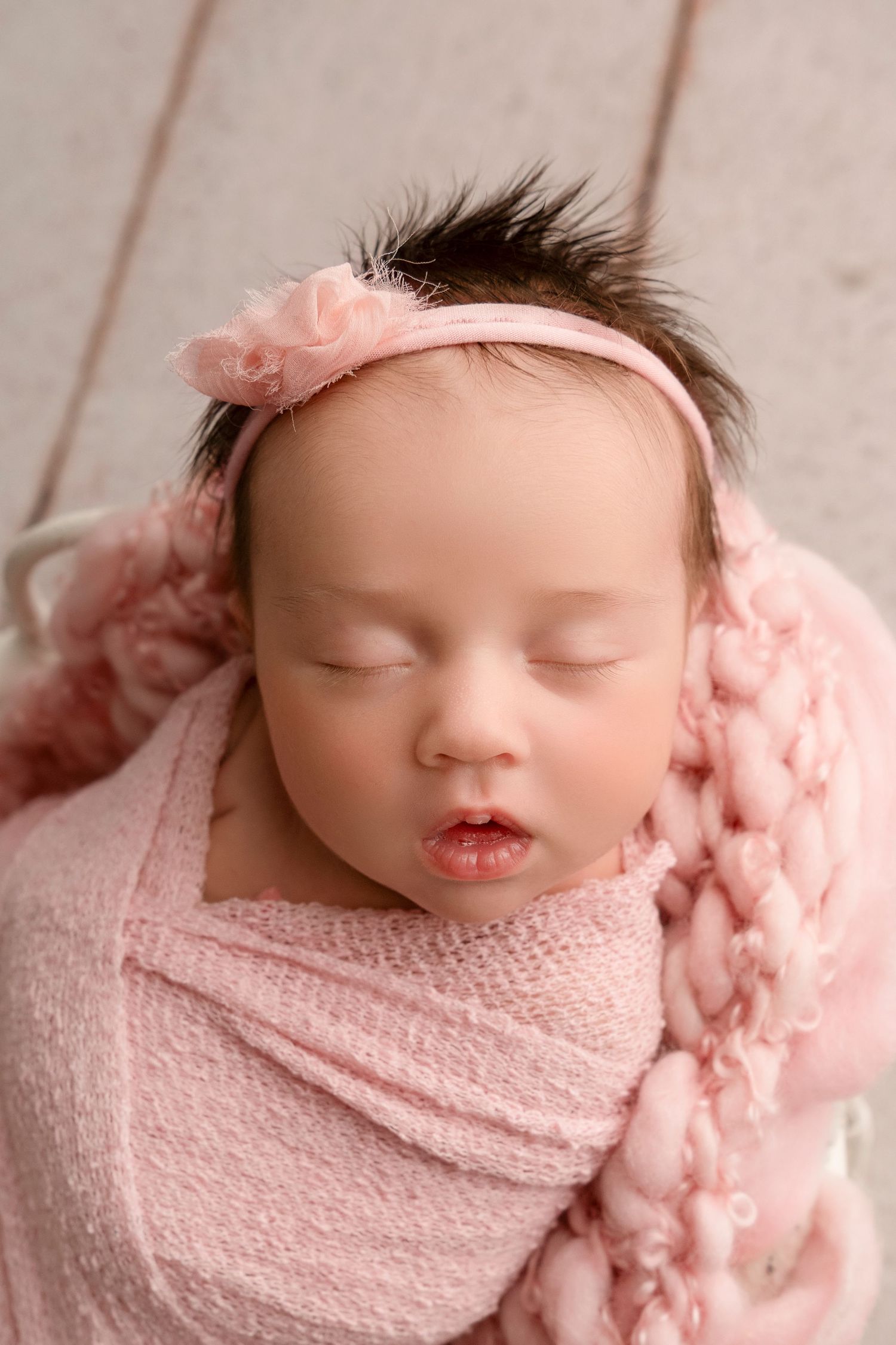 Sleeping newborn baby wrapped in soft pink blanket with matching headband in peaceful pose.