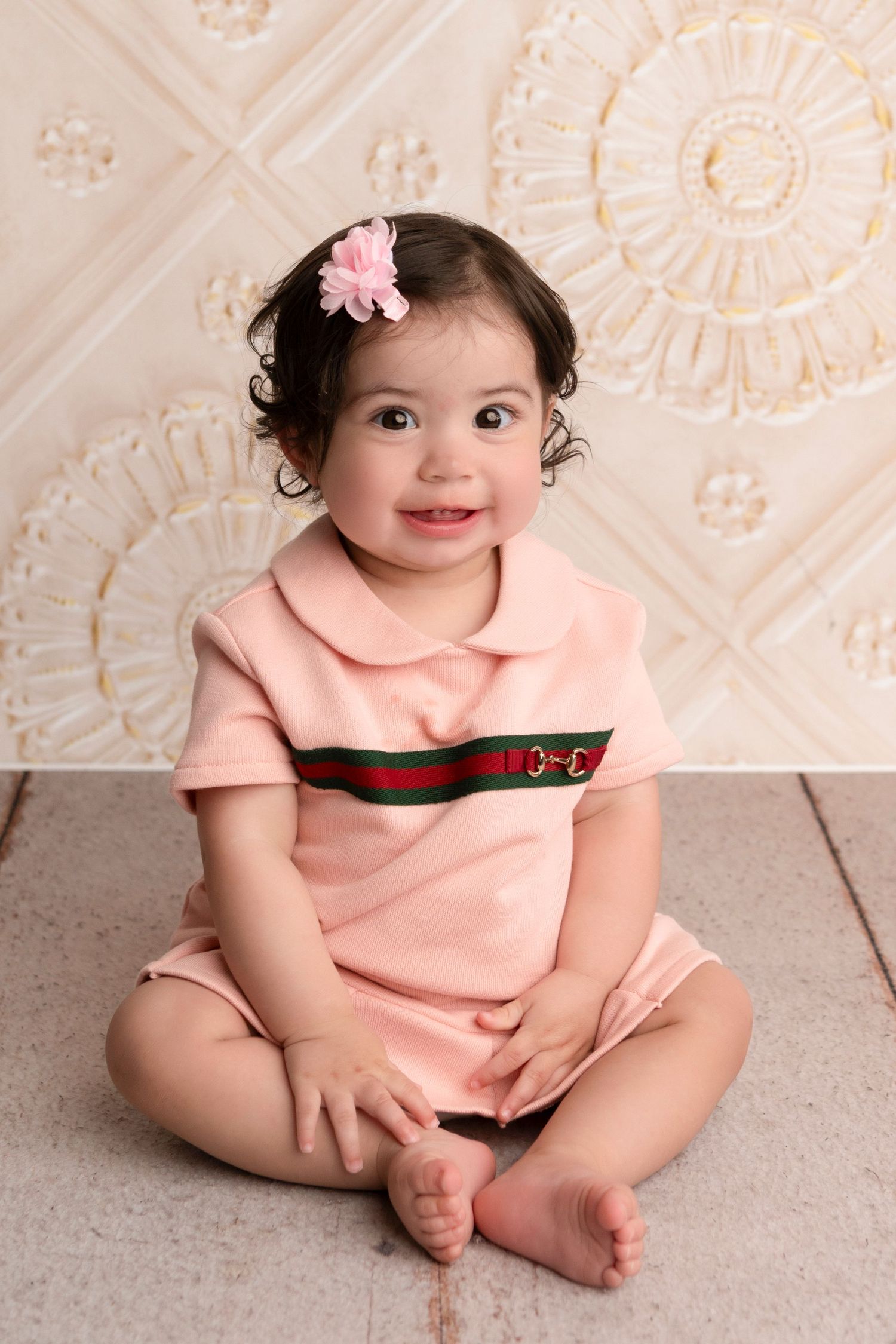 Baby wearing a peach-colored outfit sits on light-colored backdrop with ornate wall details.