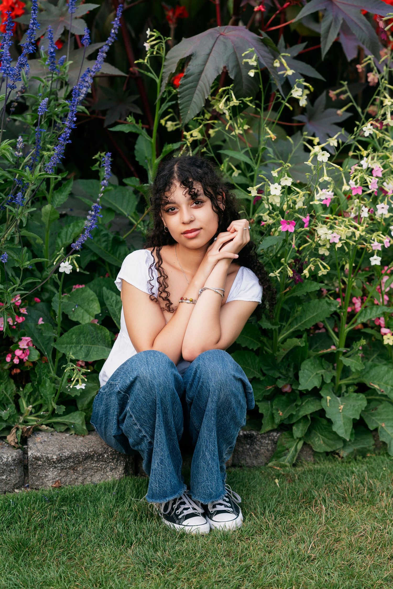A figure sits on grass amid colorful garden flowers wearing jeans and a white top with curly hair and glasses.
