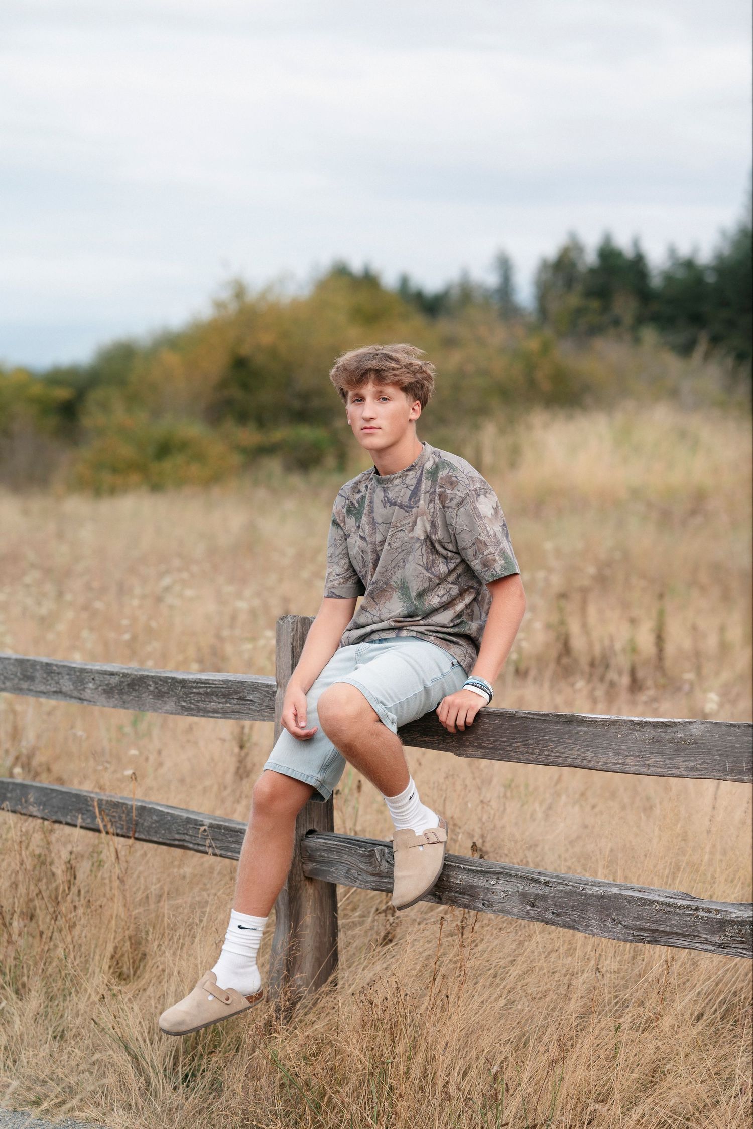Someone sits casually on a wooden fence surrounded by golden autumn grass in a rural outdoor setting.