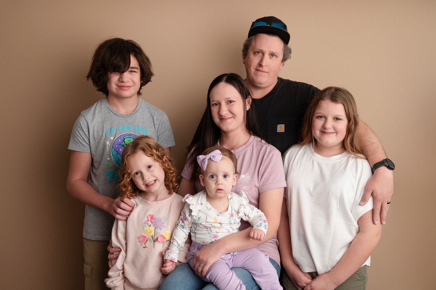 A family of six poses together, including four children and two adults, against a warm beige background in a studio portrait.