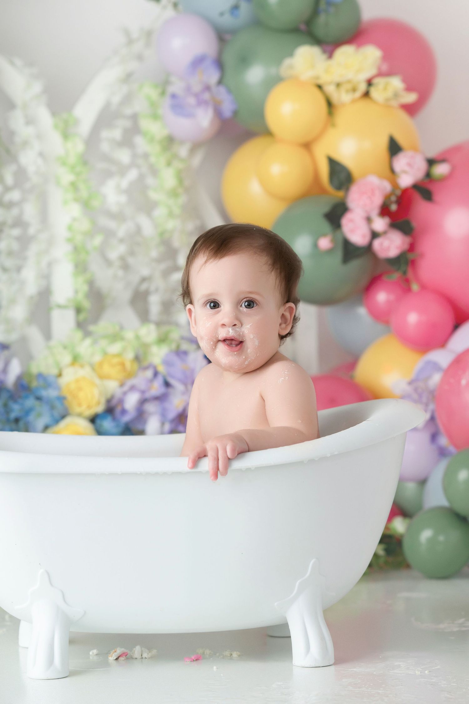 A cute baby sitting in a white mini bathtub surrounded by colorful balloons and flowers in a festive photo shoot.