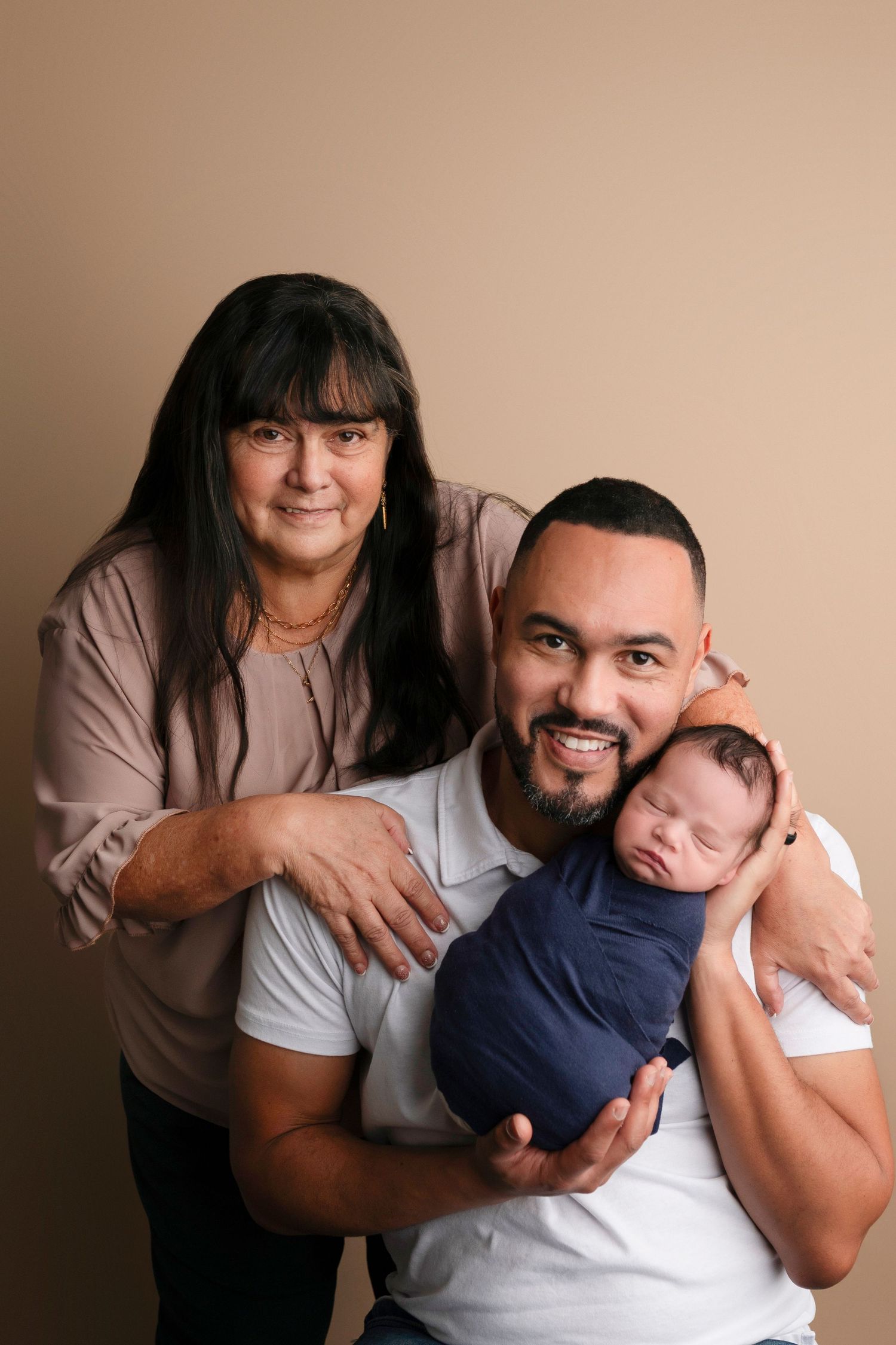 A smiling man holds a sleeping newborn wrapped in navy blue while a woman with dark hair poses beside them.