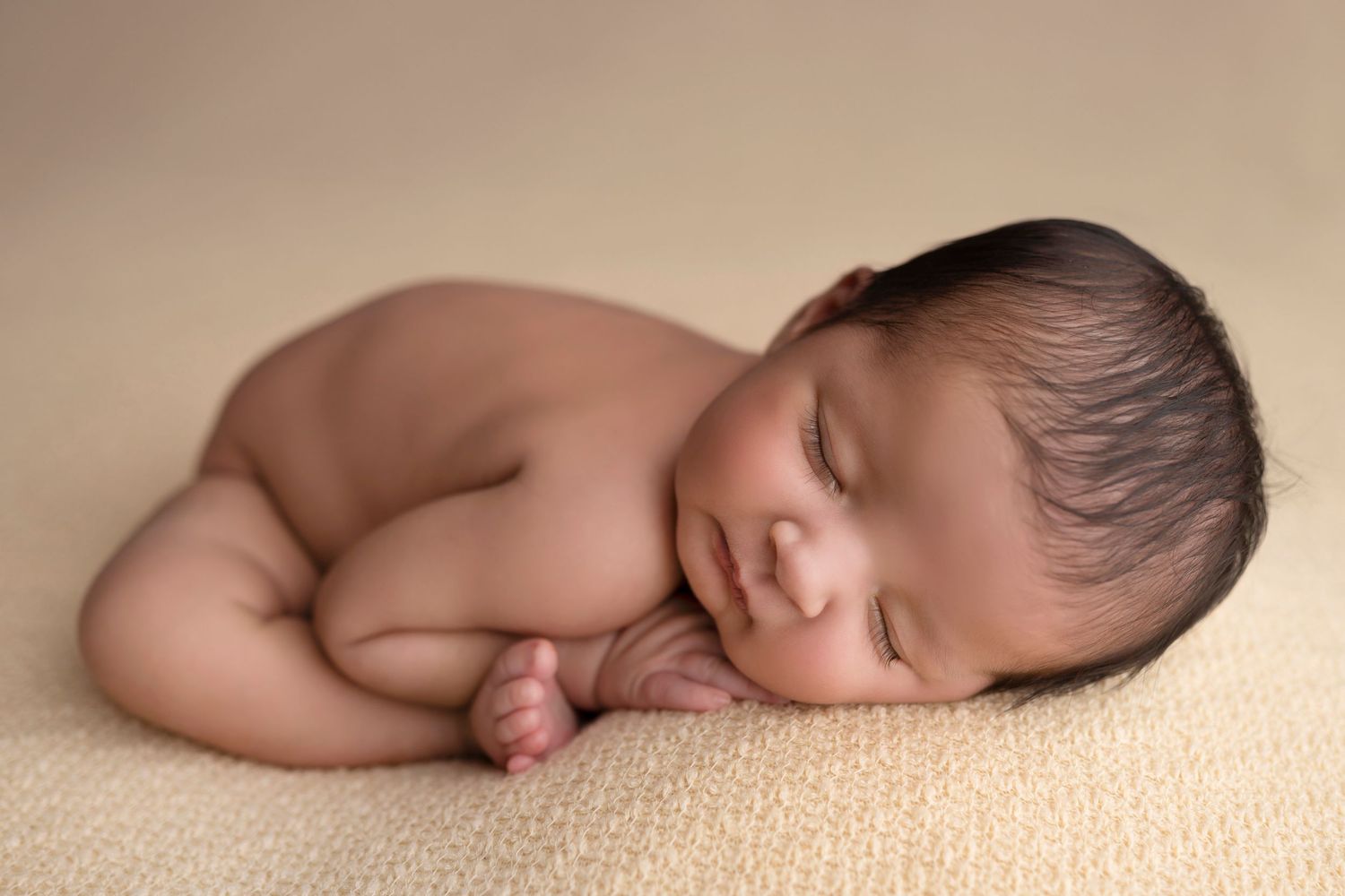 Newborn baby sleeping peacefully on soft beige blanket in natural lighting during professional photo session.