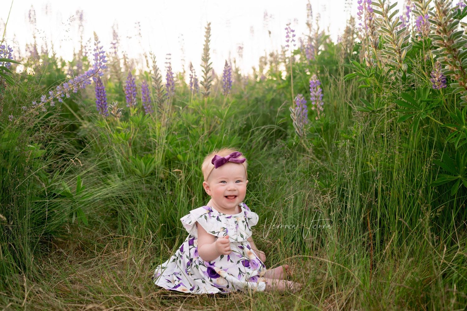 Baby girl in floral dress sits smiling in lush green field with purple lupine wildflowers during outdoor photo session.