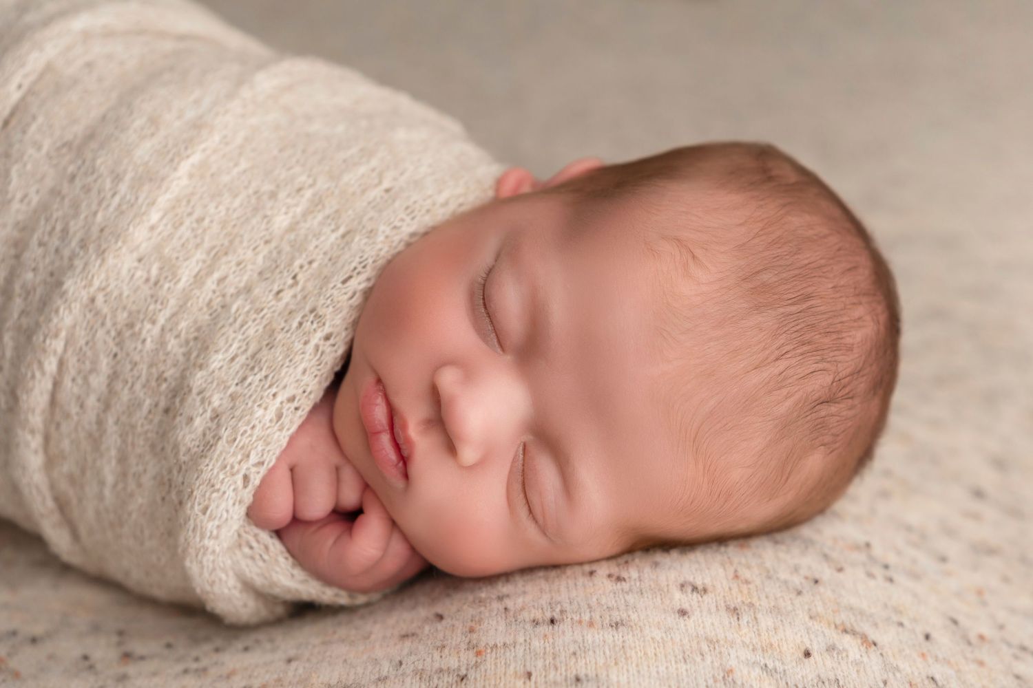 Peaceful newborn baby swaddled in soft cream wrap, sleeping soundly with tiny hands tucked under chin.