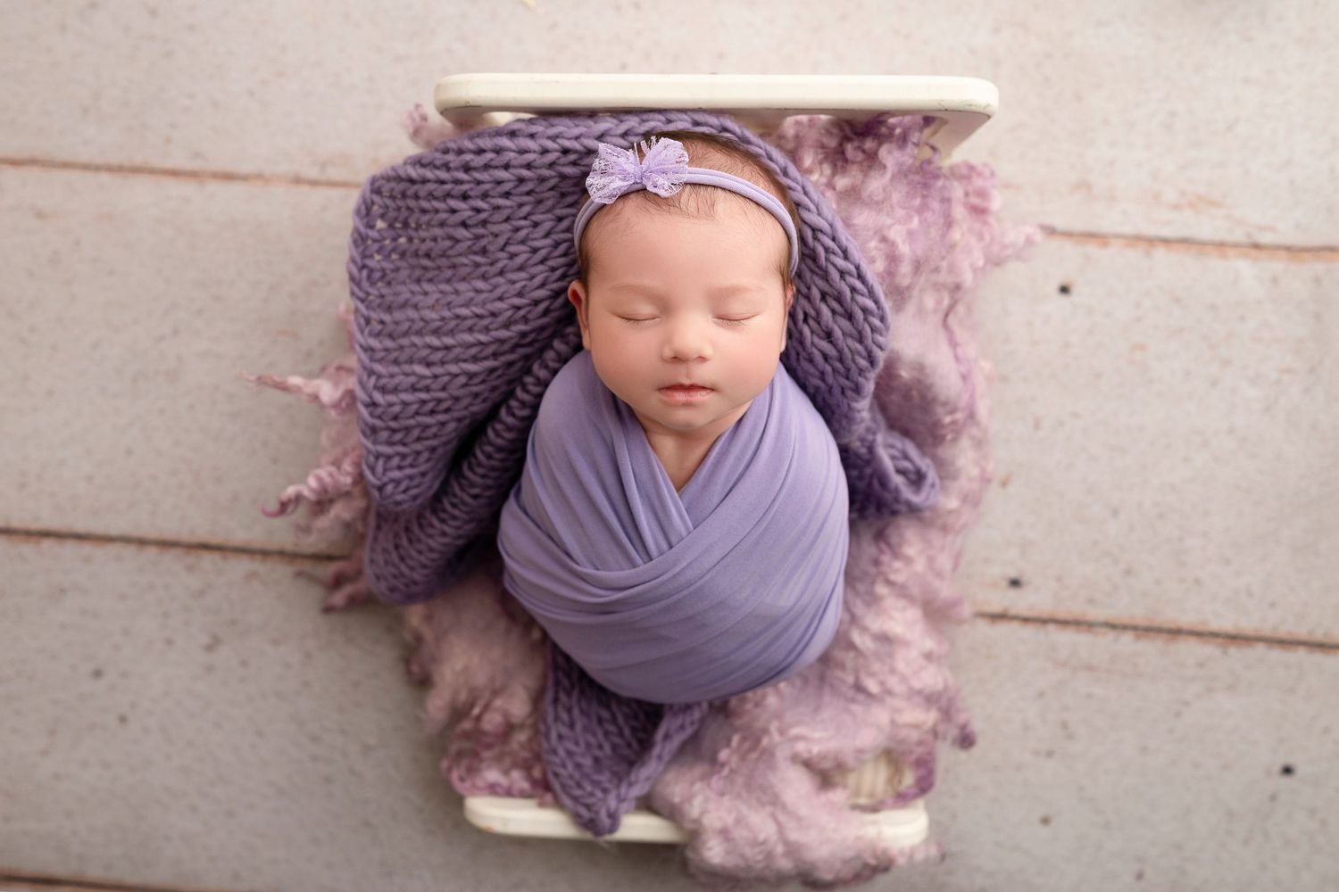 Newborn baby wrapped in purple knit swaddle with floral headband, sleeping peacefully in a white bowl prop.