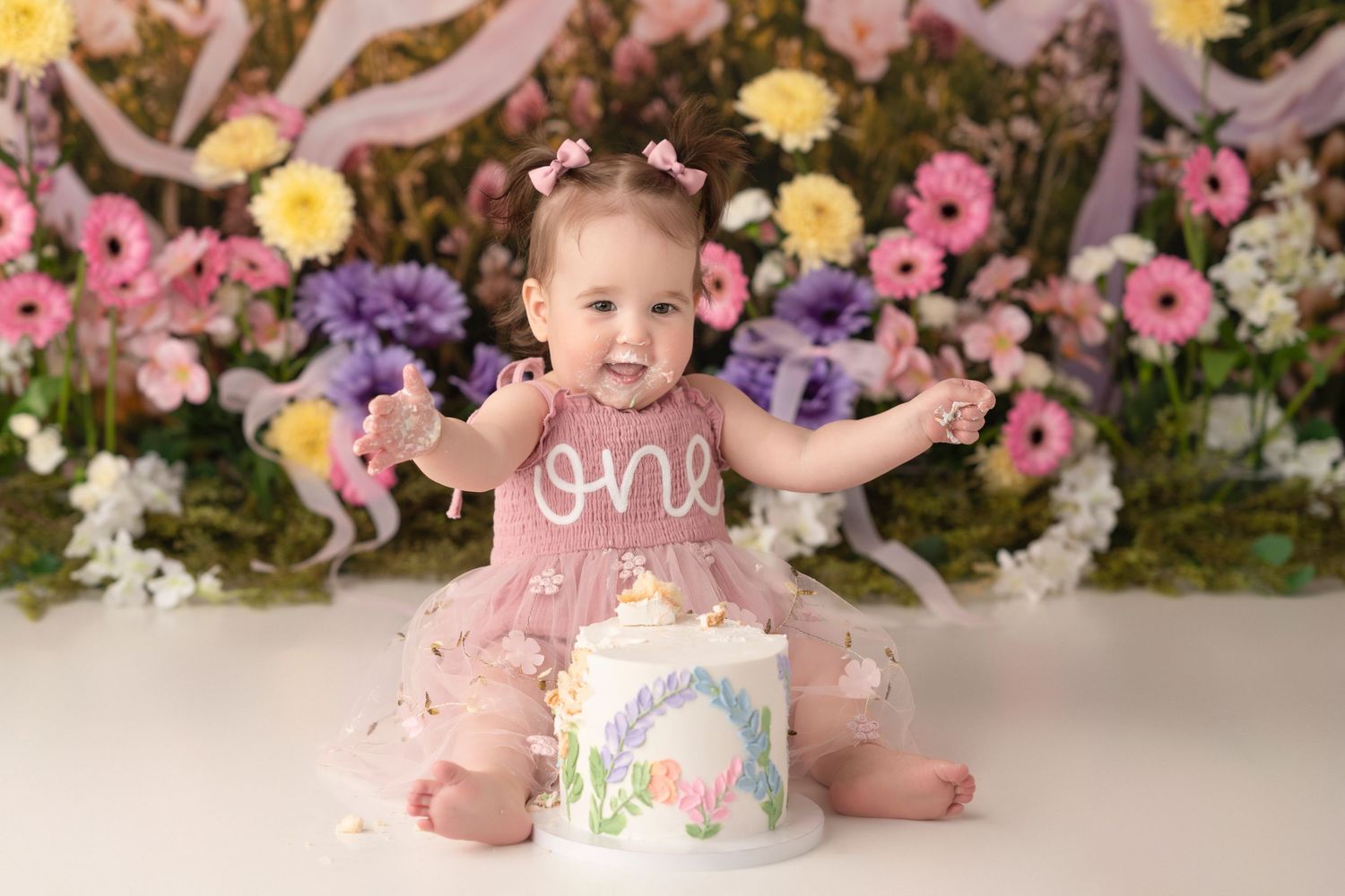 Baby girl in pink tutu dress celebrates first birthday smash cake session with floral backdrop.