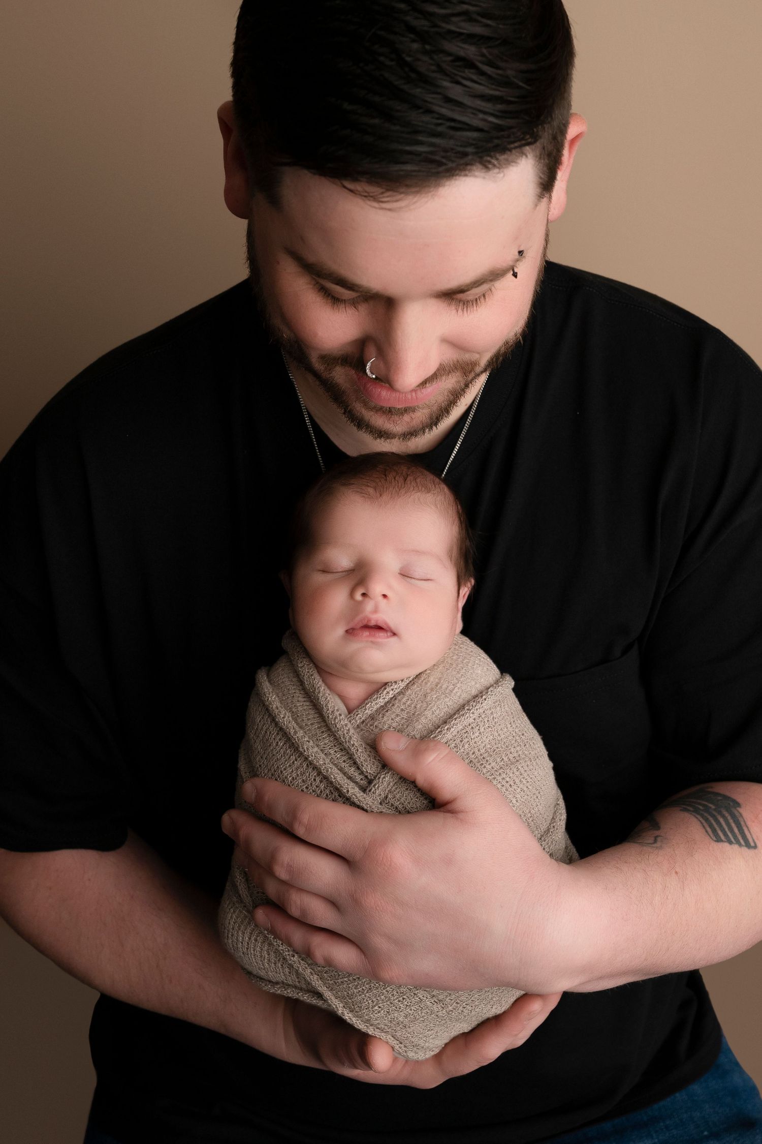 A tattooed father in a black shirt tenderly holds his swaddled sleeping newborn baby against his chest.