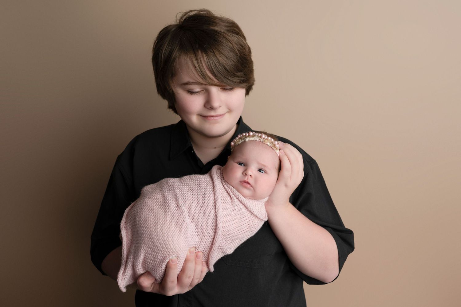 Older child tenderly holds swaddled newborn baby in pink wrap with floral headband against beige backdrop.