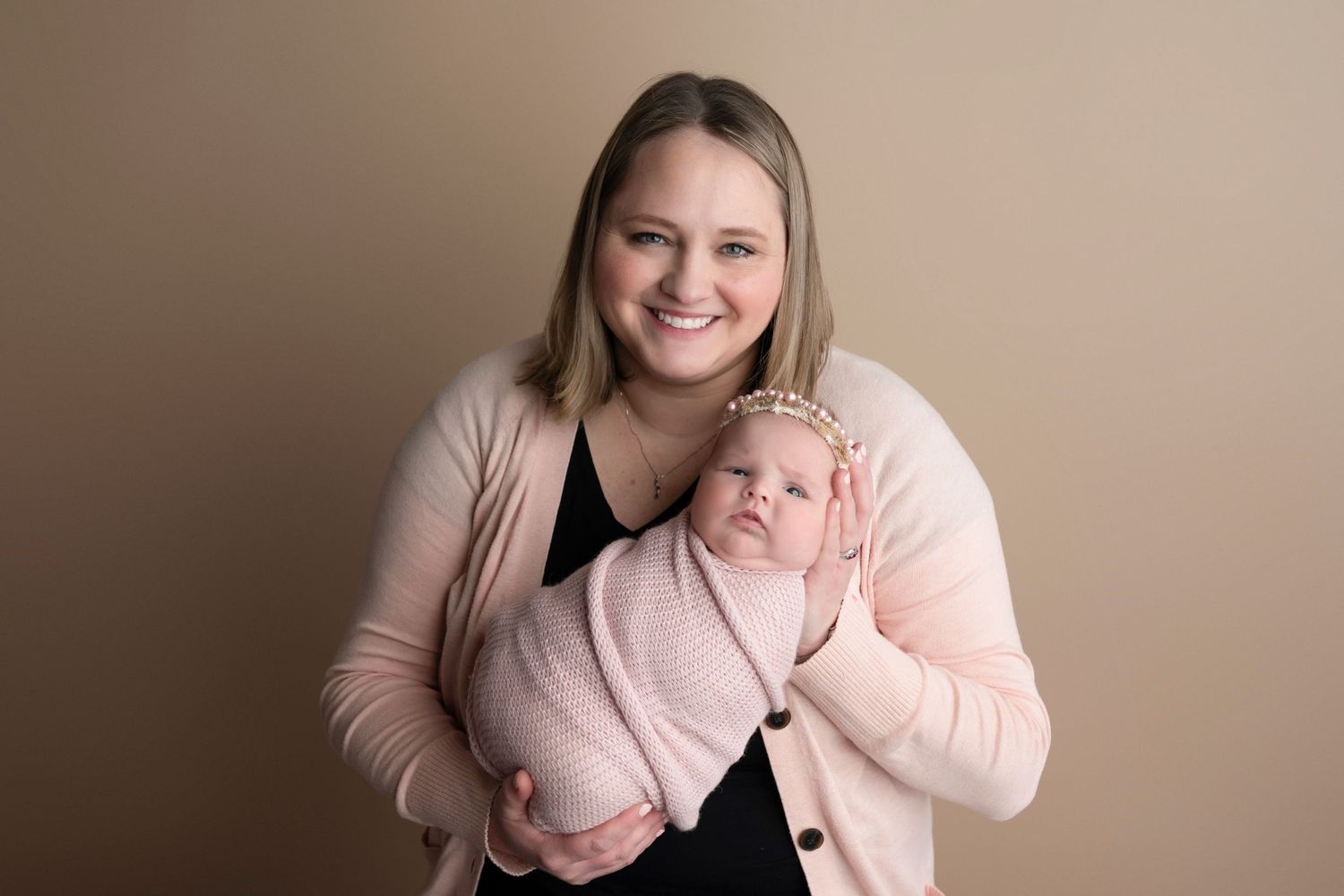 A smiling mother in a pink cardigan holds her swaddled newborn baby with a floral headband against a beige backdrop.