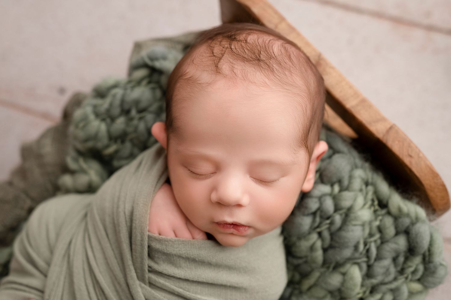 Newborn baby sleeping peacefully on sage green textured blanket in rustic wooden bed.