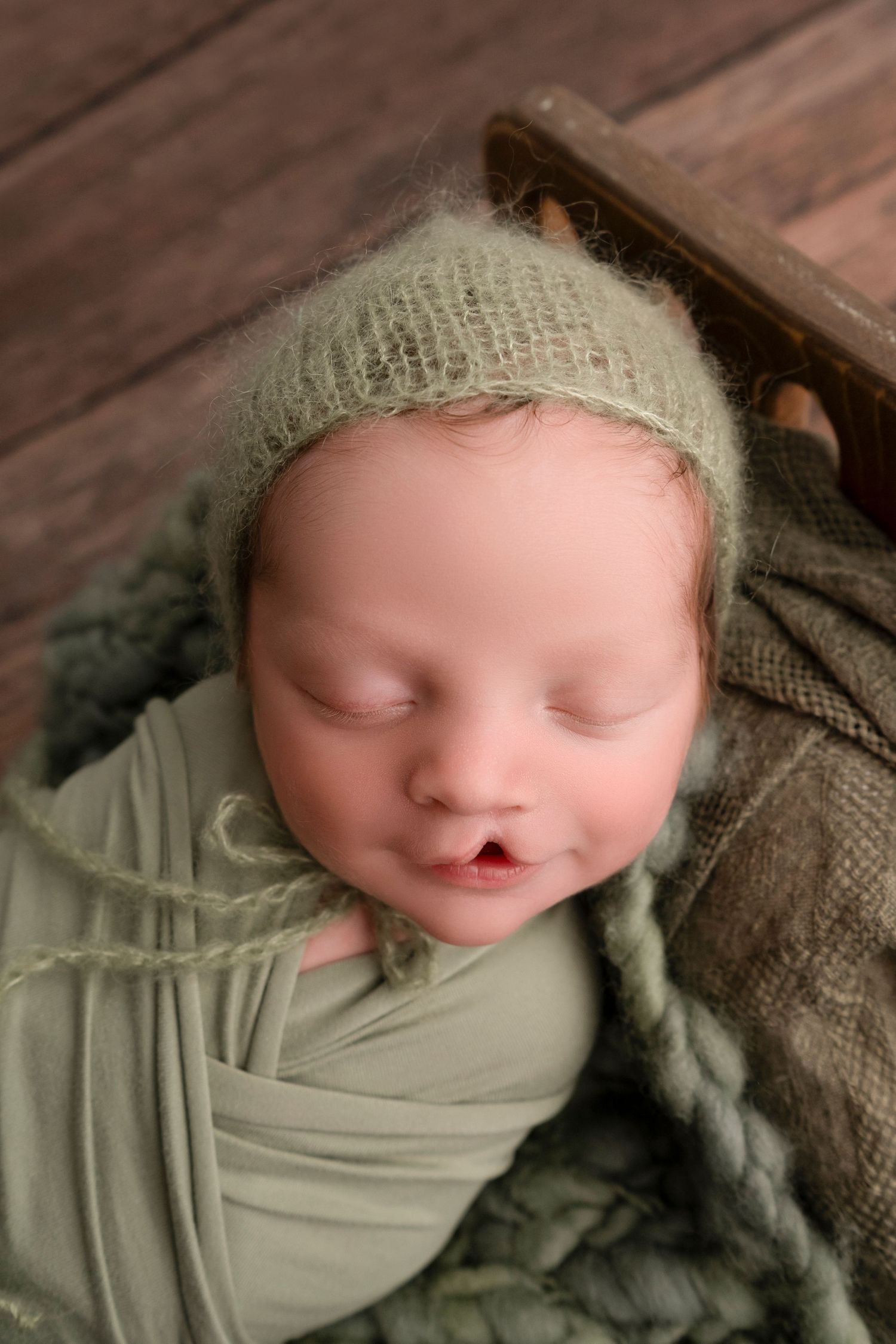 A sleeping newborn wearing a mint green wrap and knit bonnet lies peacefully under a cozy blanket.