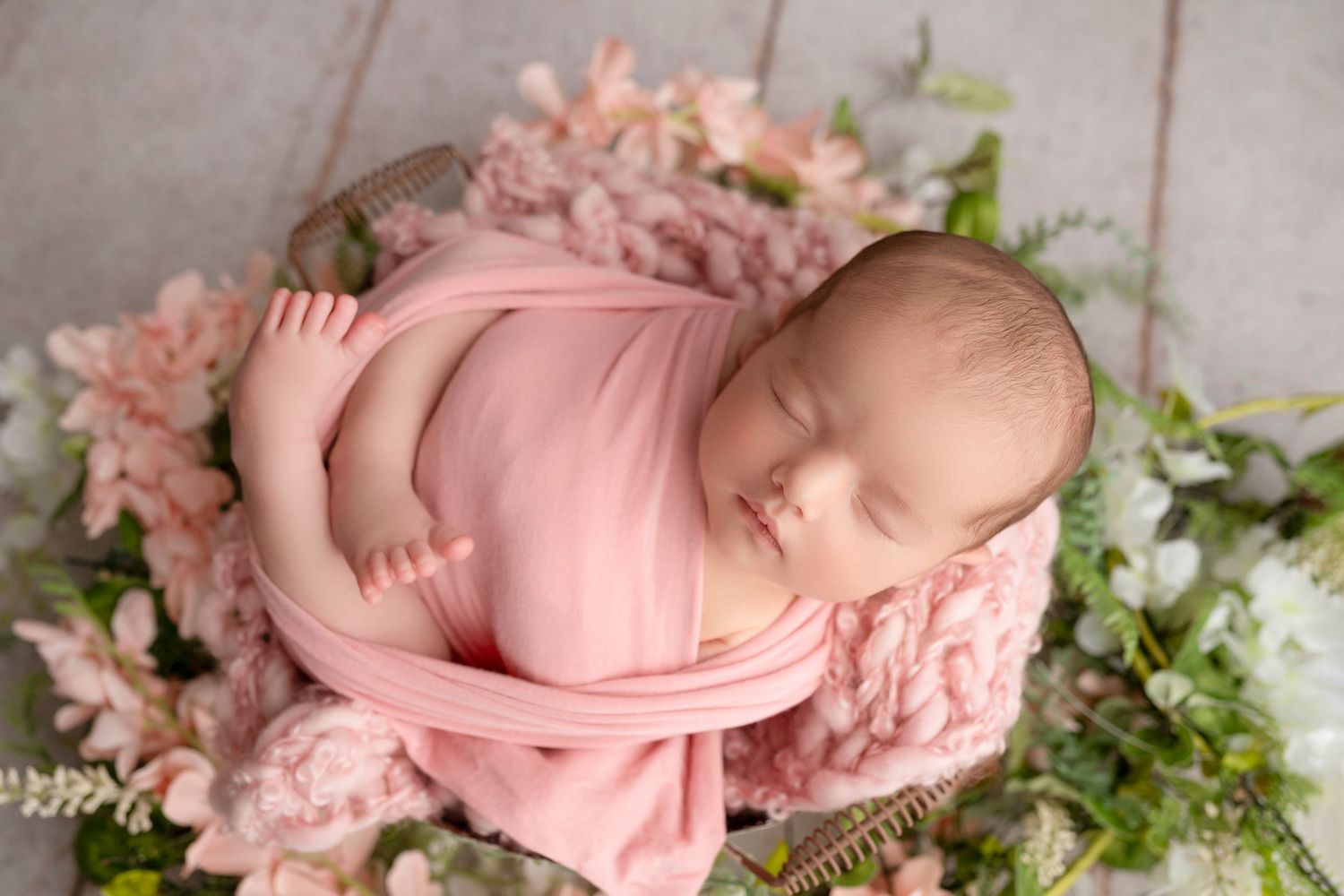 Newborn baby sleeping peacefully on pink blanket surrounded by delicate white flowers and greenery.