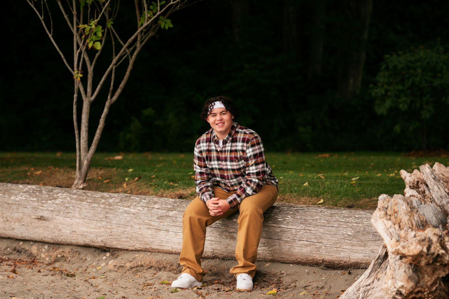 Person in plaid shirt and brown pants sits on rustic wooden log bench in outdoor forest setting.