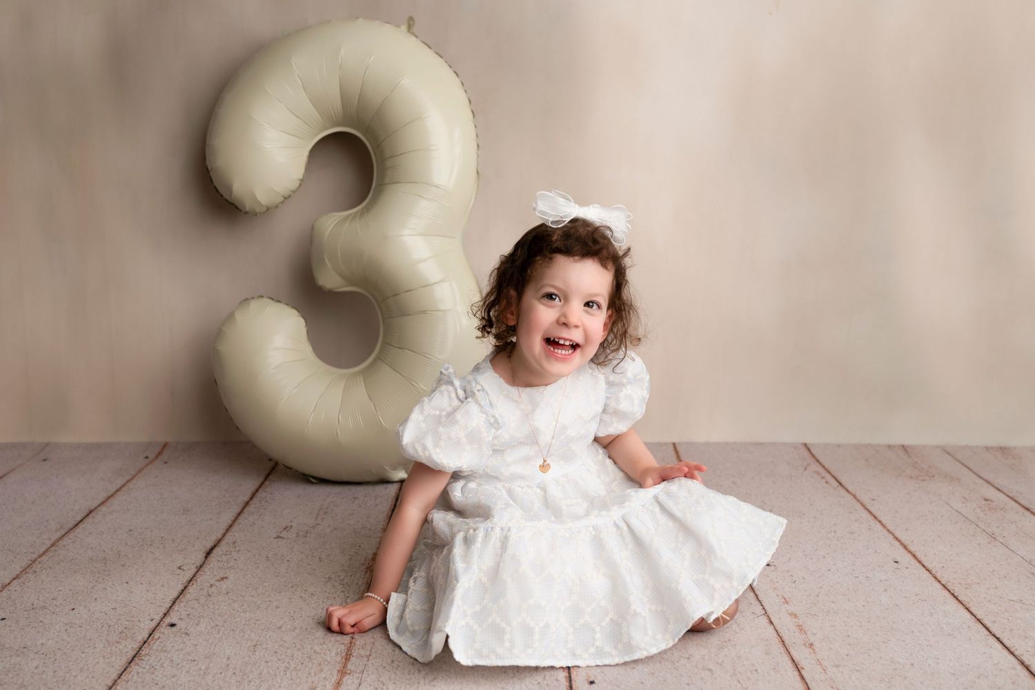 A child in a white dress sits next to a large number 3 prop on a wooden floor for a photo session.