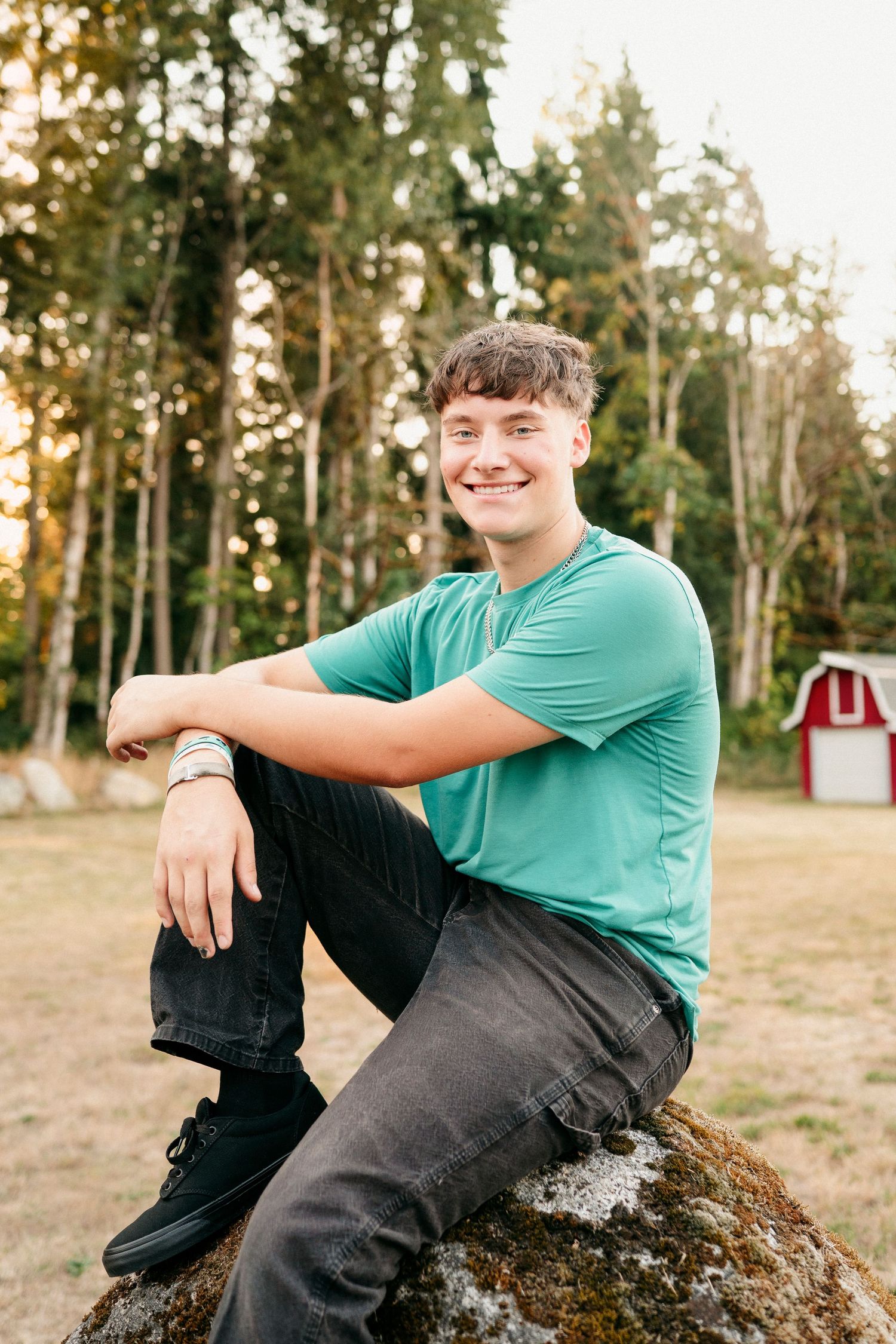 Person in teal t-shirt and dark pants sits casually on a rock outdoors with trees and red barn in background.