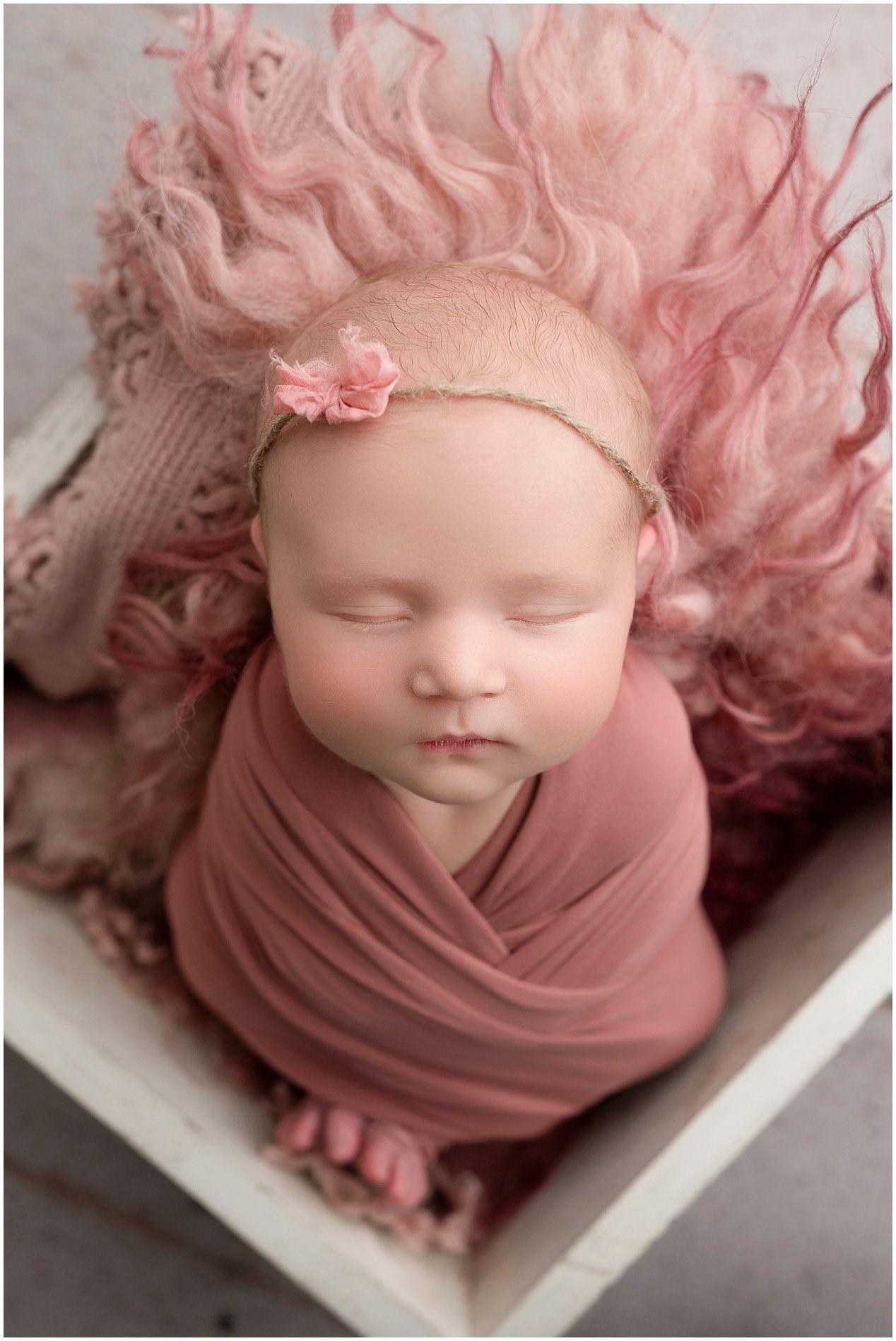 A sleeping newborn baby wrapped in dusty rose fabric with pink curly hair decoration against white backdrop.