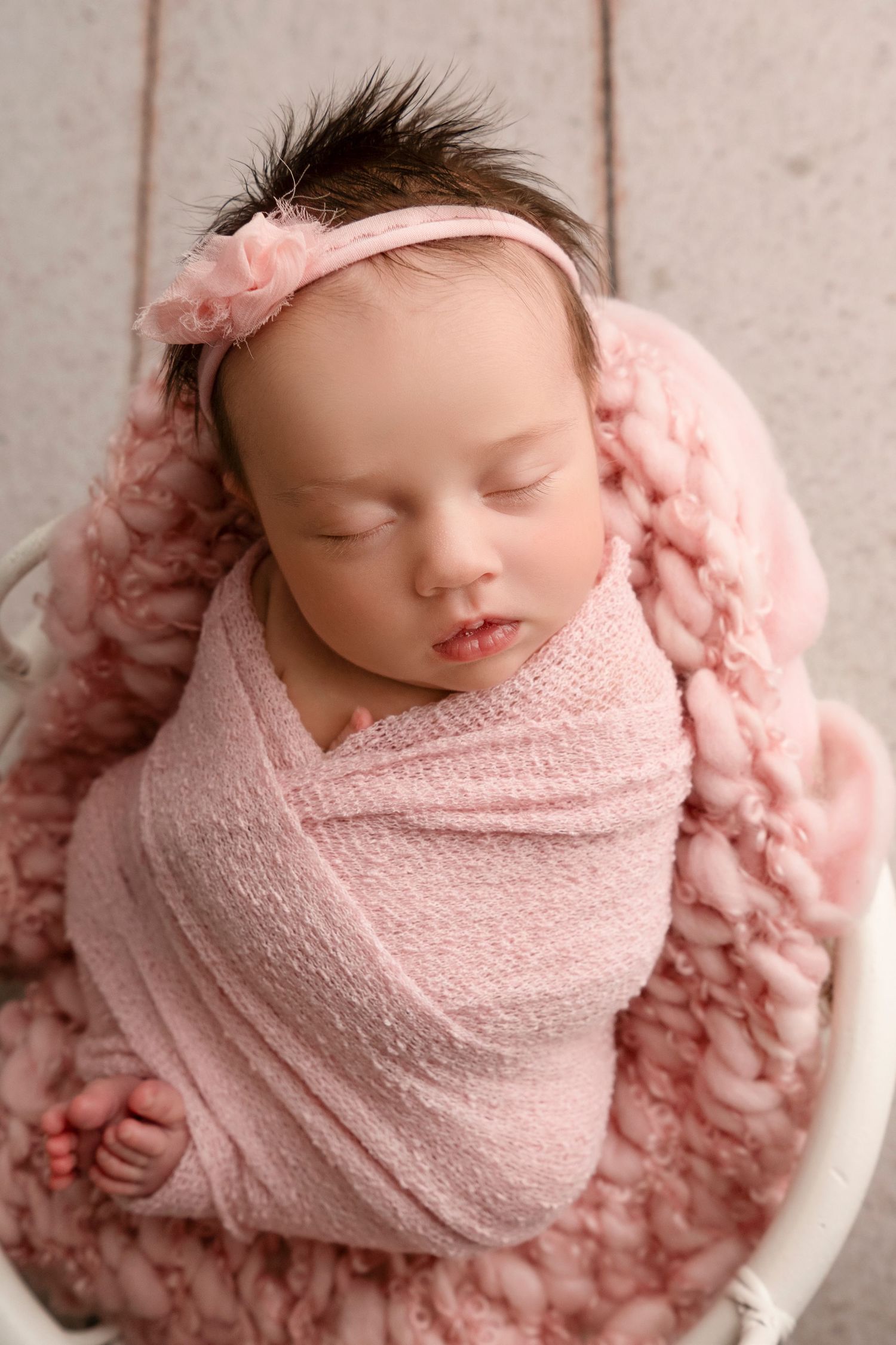 A sleeping newborn swaddled in soft pink fabric rests peacefully in a white bowl against a pink textured backdrop.