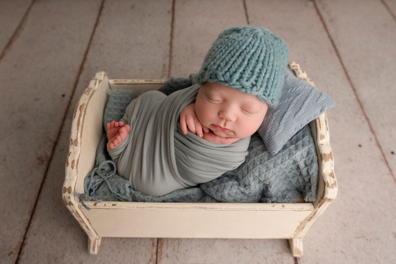 A sleeping newborn wearing a teal knit outfit rests peacefully in a rustic wooden crate on a pink floor.