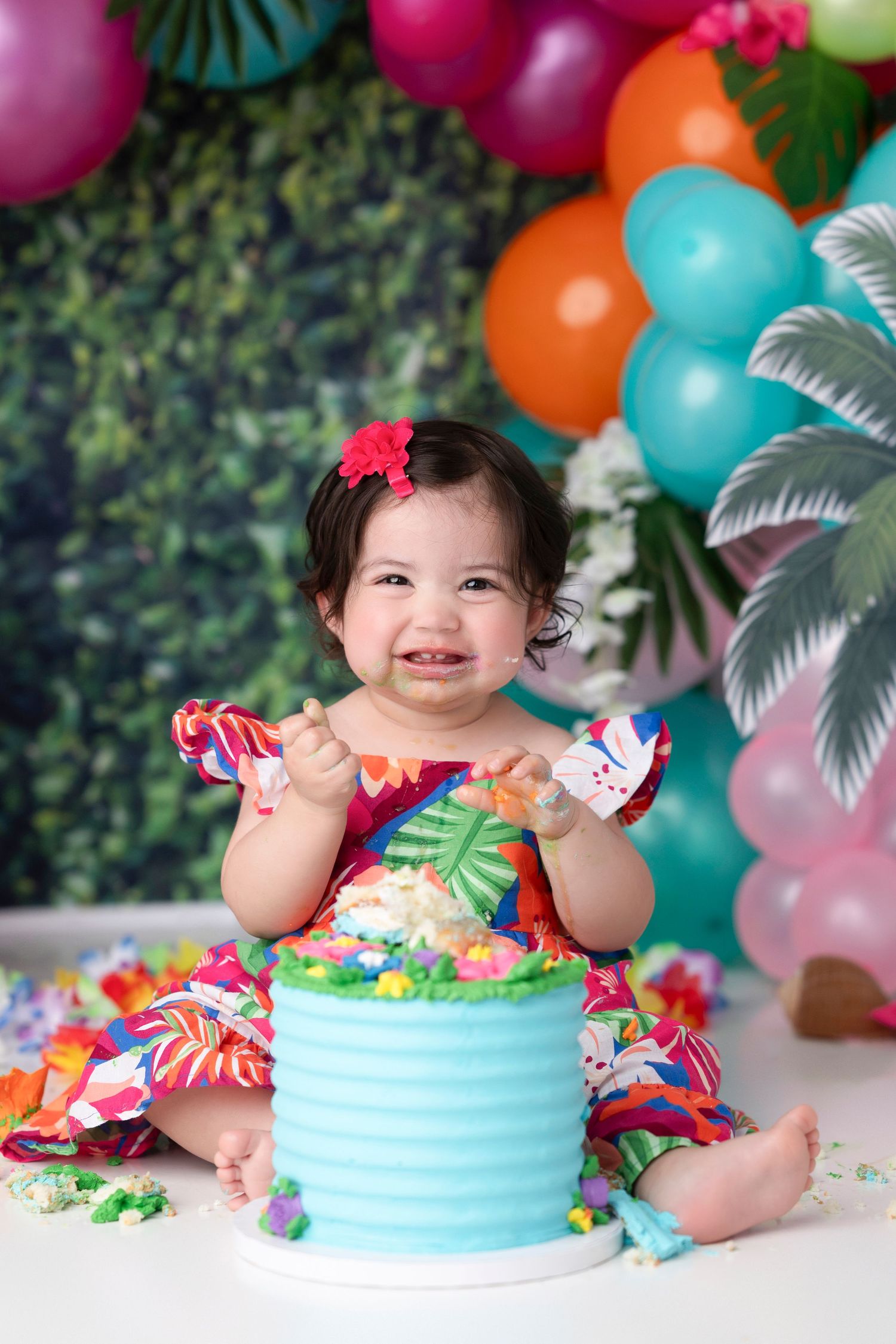 A festive cake smash photo with colorful balloons and a blue buttercream cake against a tropical backdrop.