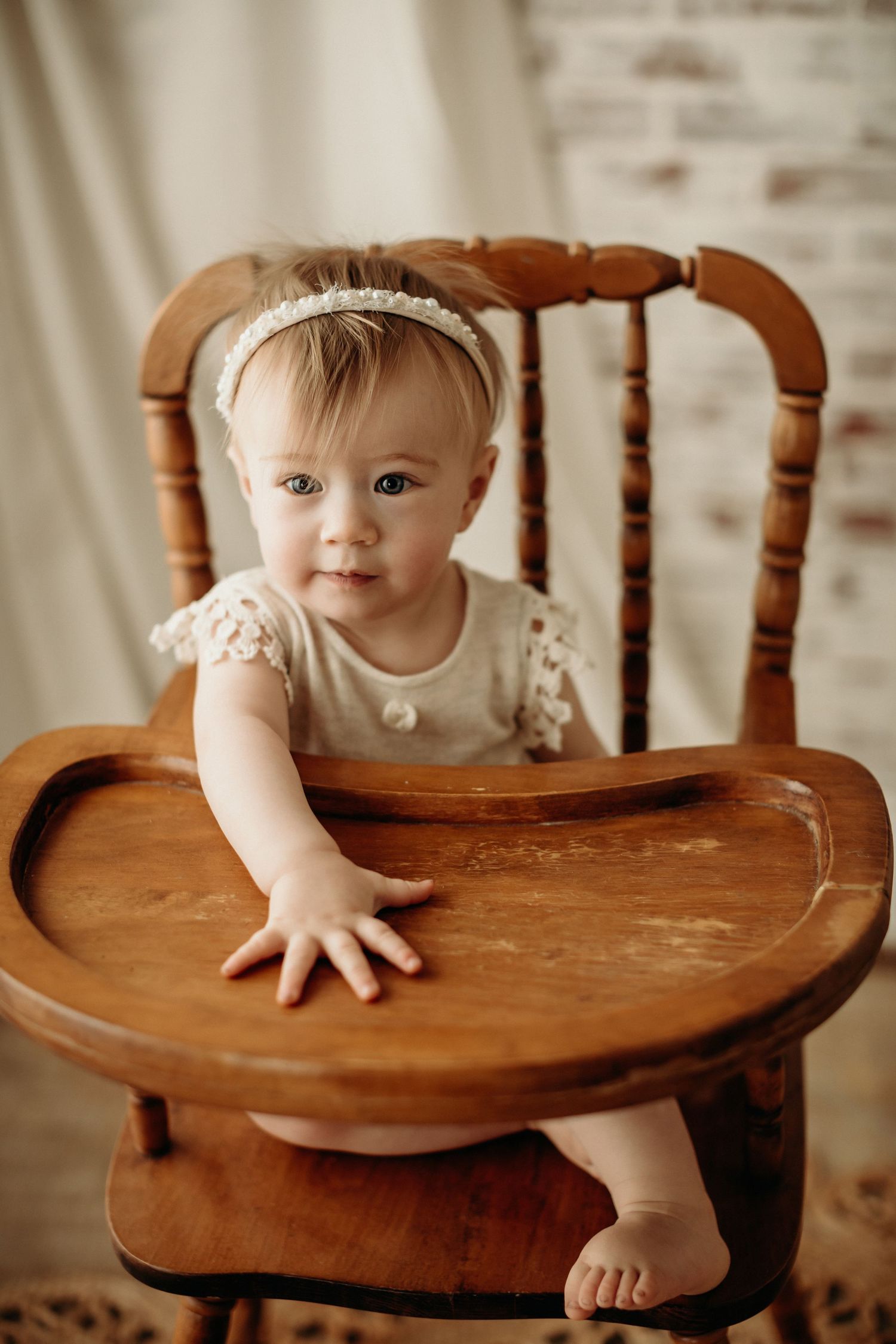 A small child sits in a vintage wooden high chair wearing a cream-colored dress with lace details.