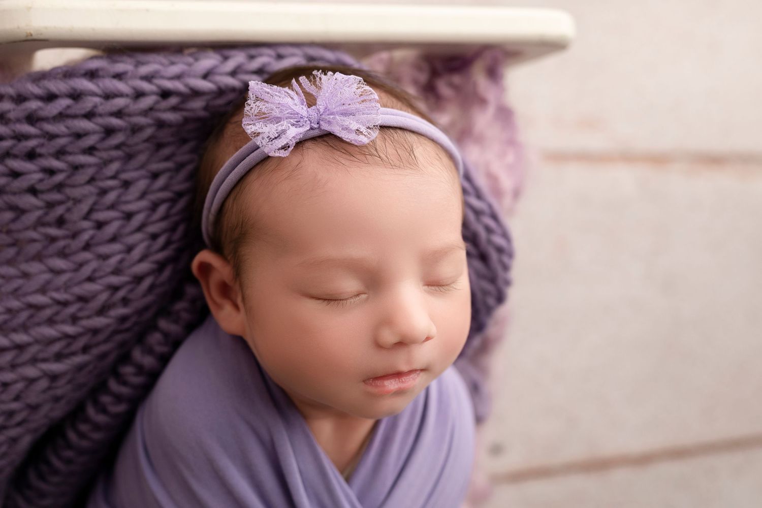 Sleeping newborn baby girl wrapped in purple knit blanket wearing a lavender bow headband.