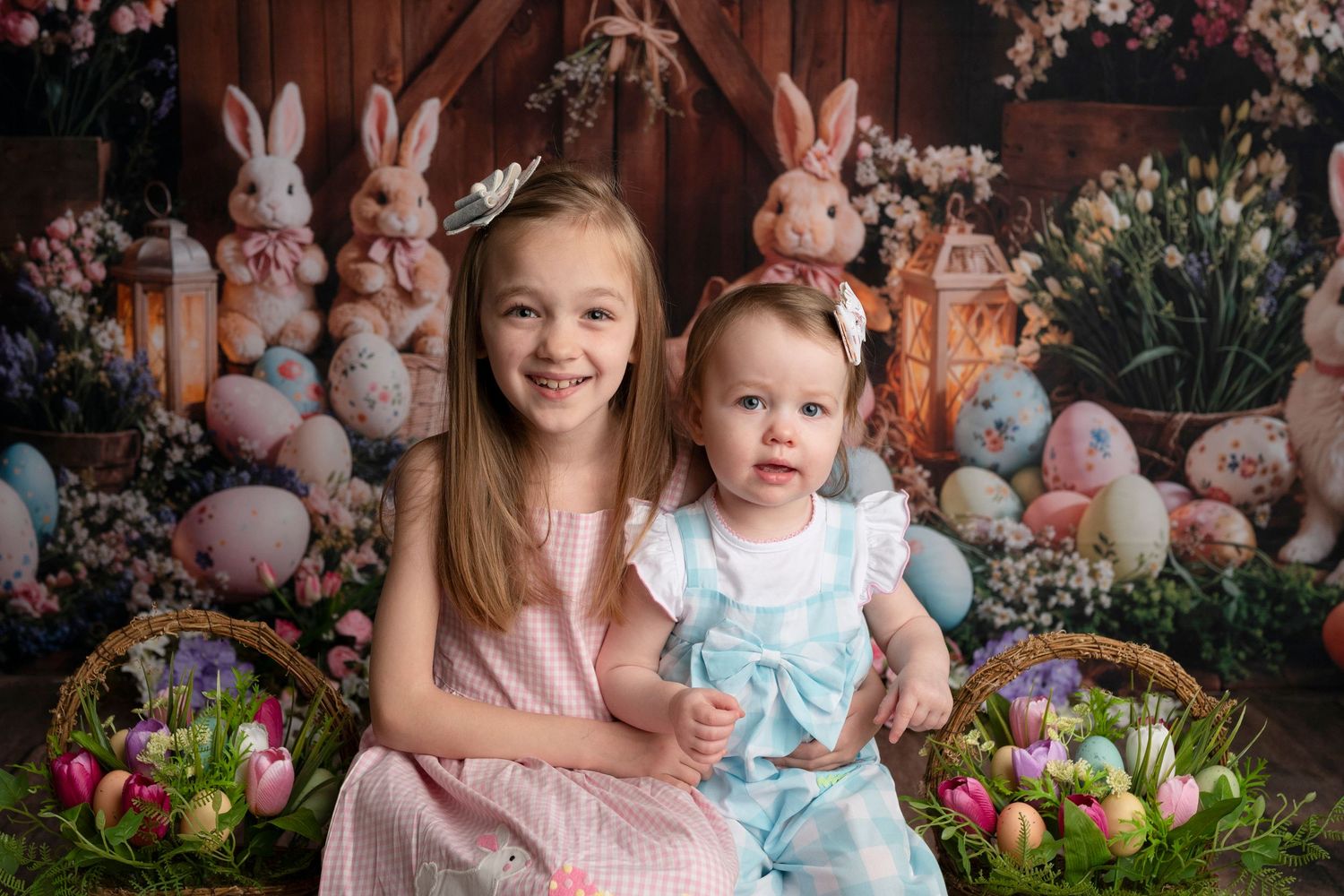Two girls in pastel dresses pose with Easter eggs, bunnies, and flower baskets in a festive spring photo.