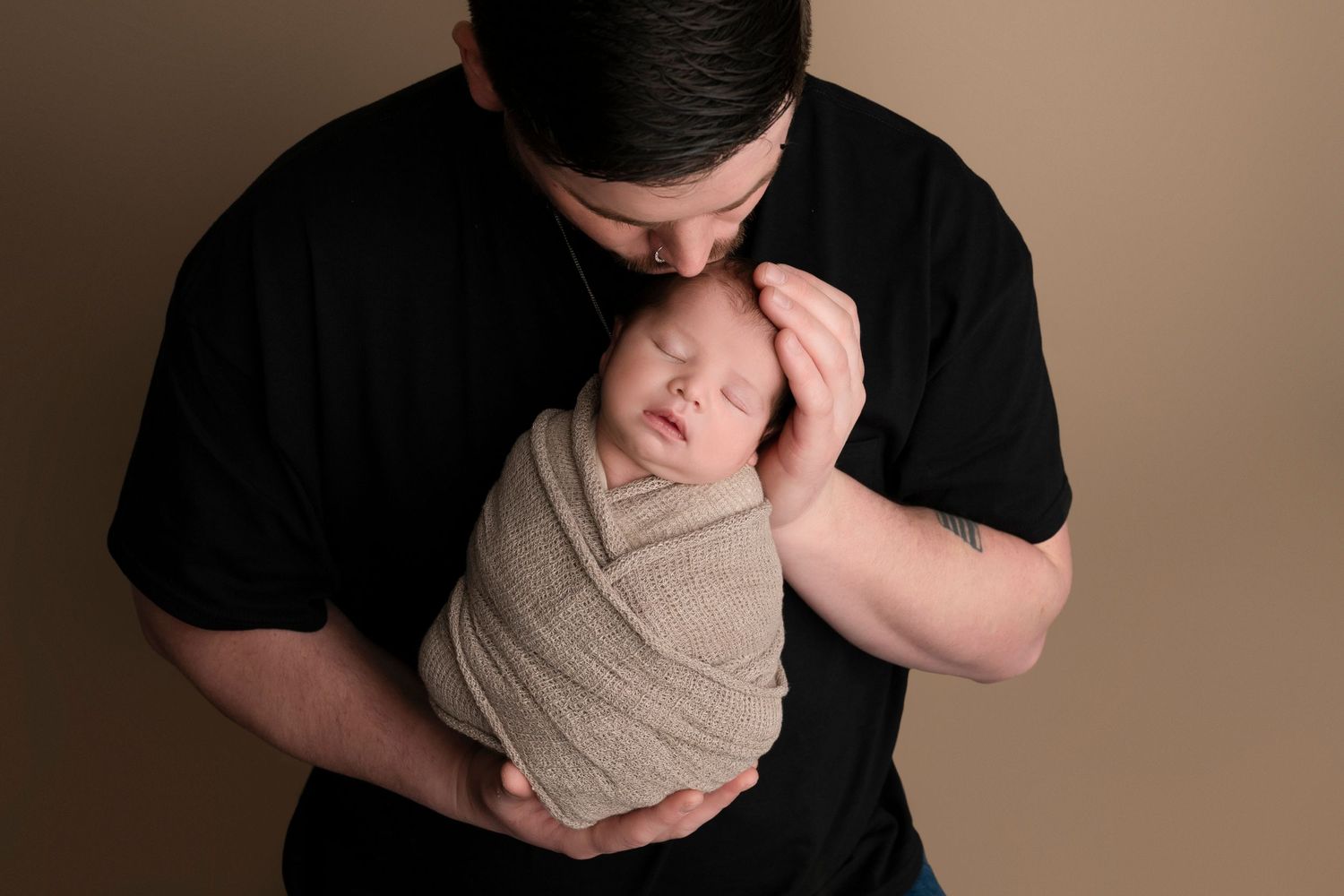 Father in black shirt kissing sleeping newborn baby wrapped in beige swaddle against neutral background.