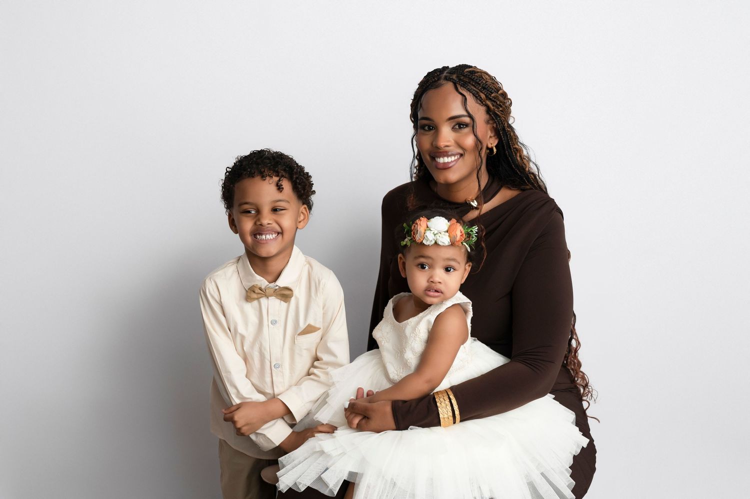 A smiling woman in brown holds a baby girl in white dress while a young boy in bow tie poses beside them.