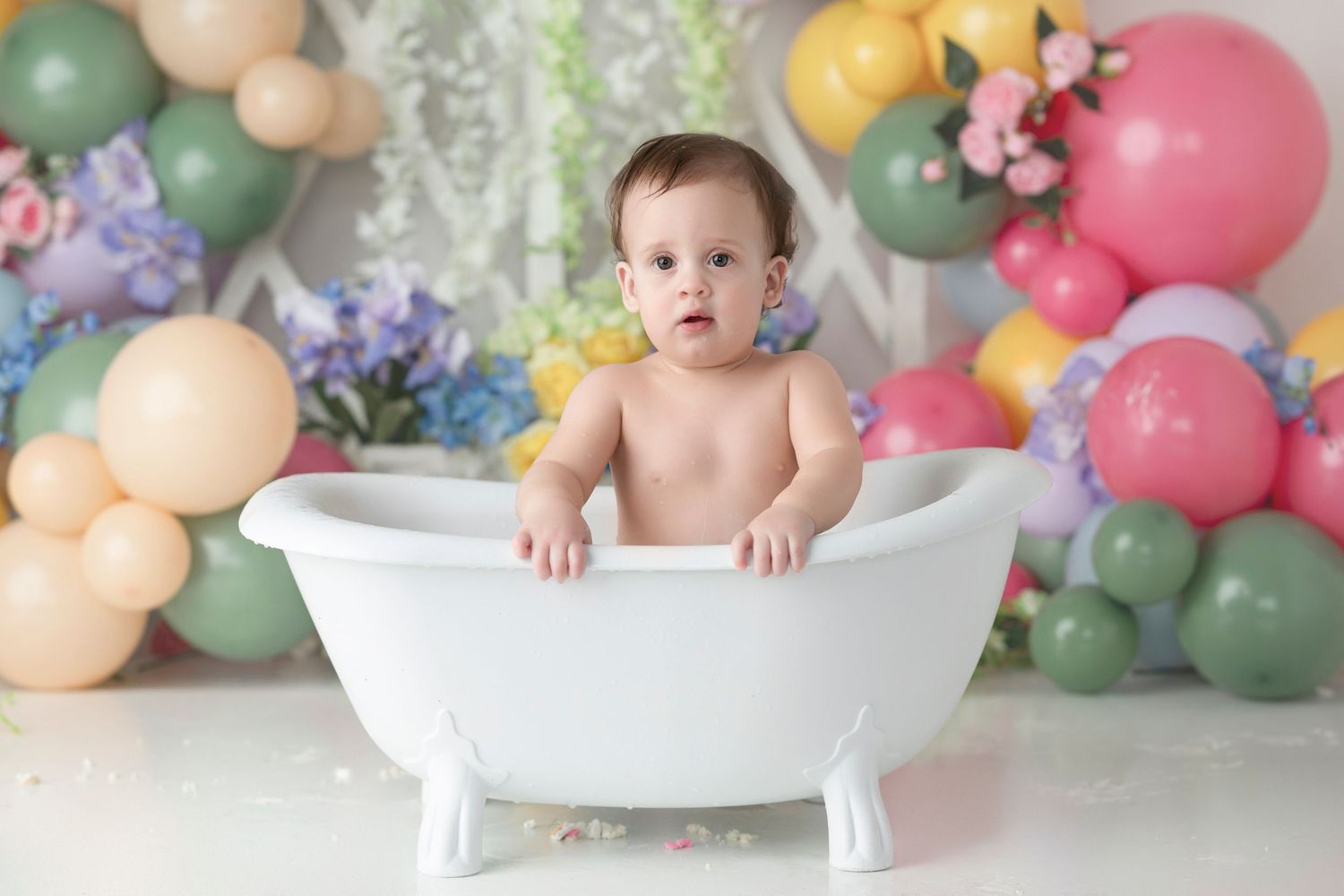 Baby sitting in white mini bathtub surrounded by colorful balloons and flowers in a festive first birthday photo session.