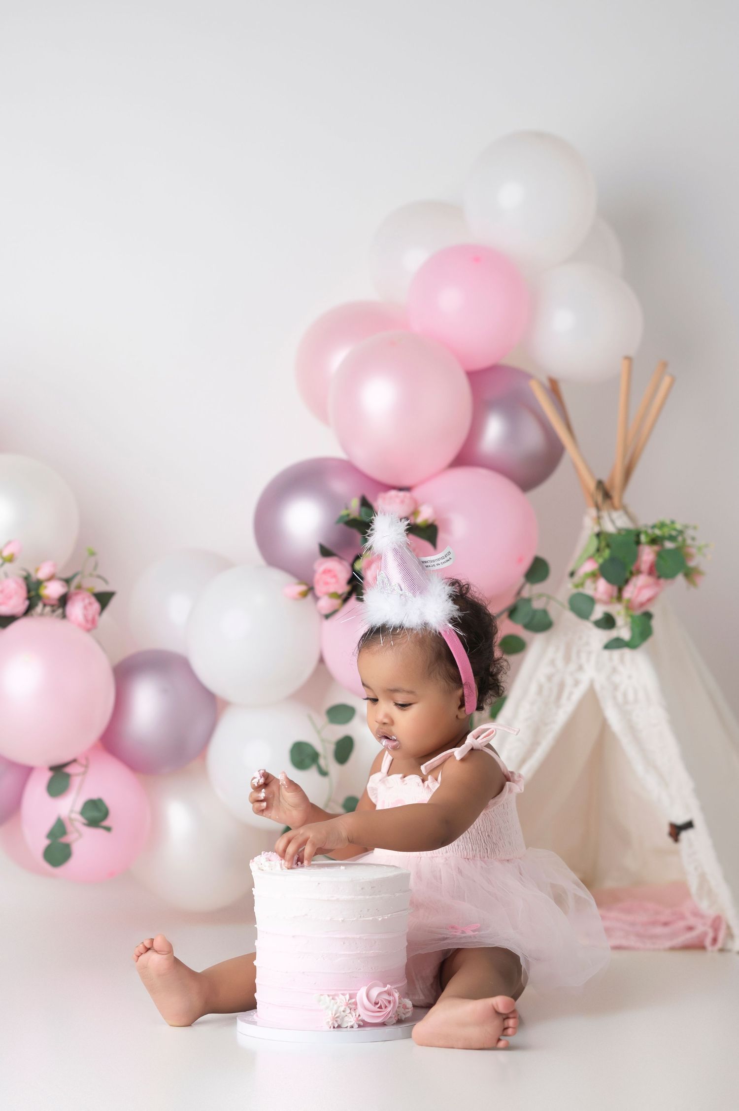 Baby girl in pink tutu touches first birthday cake surrounded by pink and white balloons and a boho teepee backdrop.