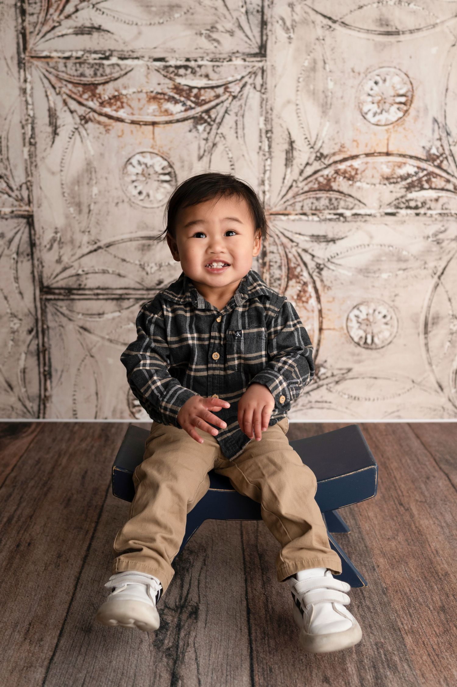 A smiling toddler boy in a plaid shirt and khaki pants sits on a navy box against a vintage ornate backdrop.