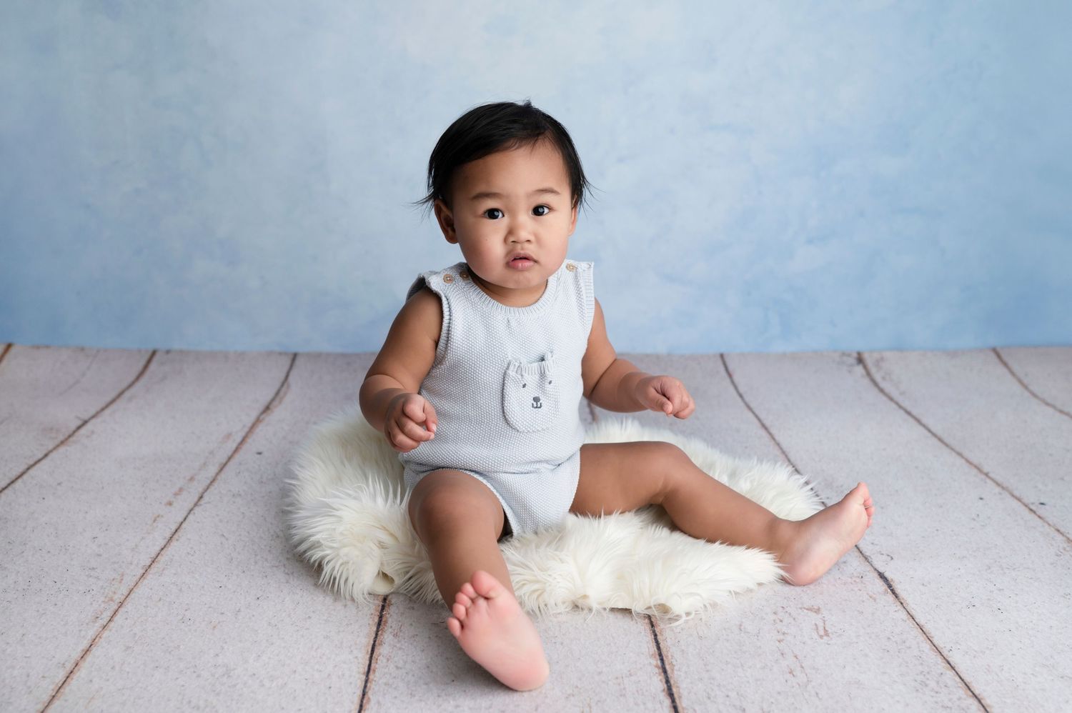 Adorable baby girl sitting on fluffy white rug in light blue romper against soft blue backdrop.