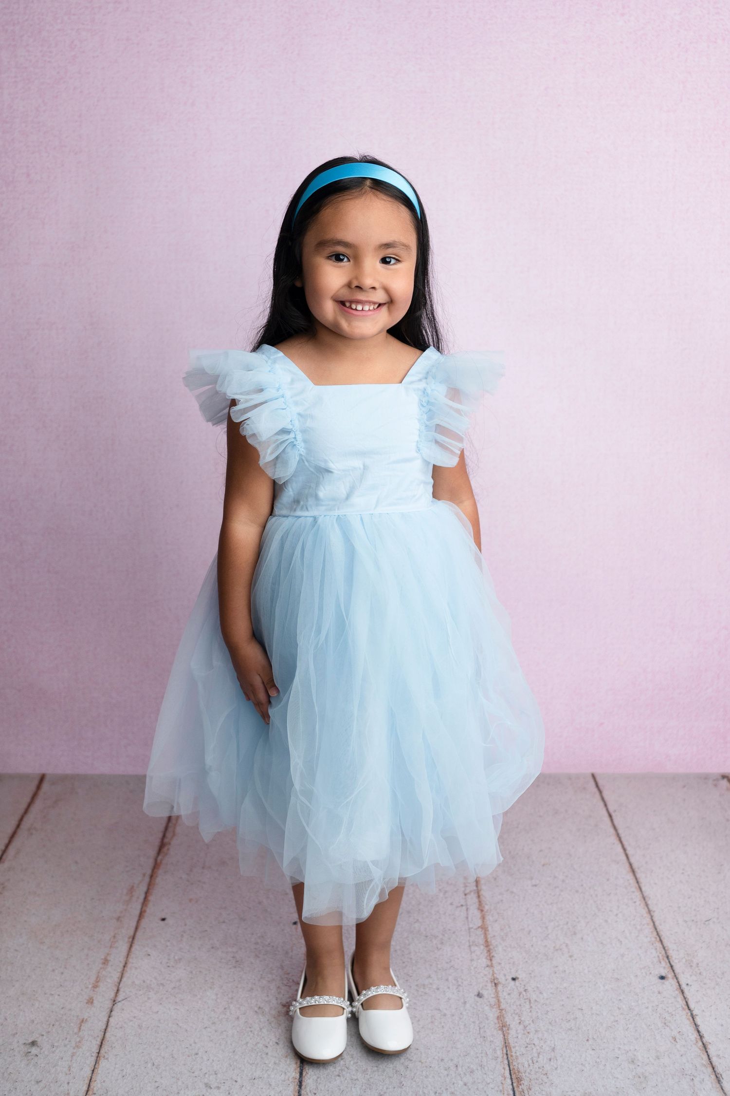 Young girl smiling in a light blue tulle dress with flutter sleeves and matching headband, posing against a pink background.