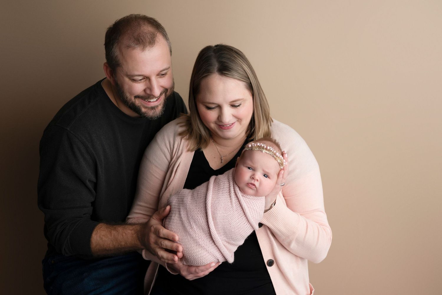 Happy parents smiling at their swaddled newborn baby in a pink wrap during a professional studio portrait session.
