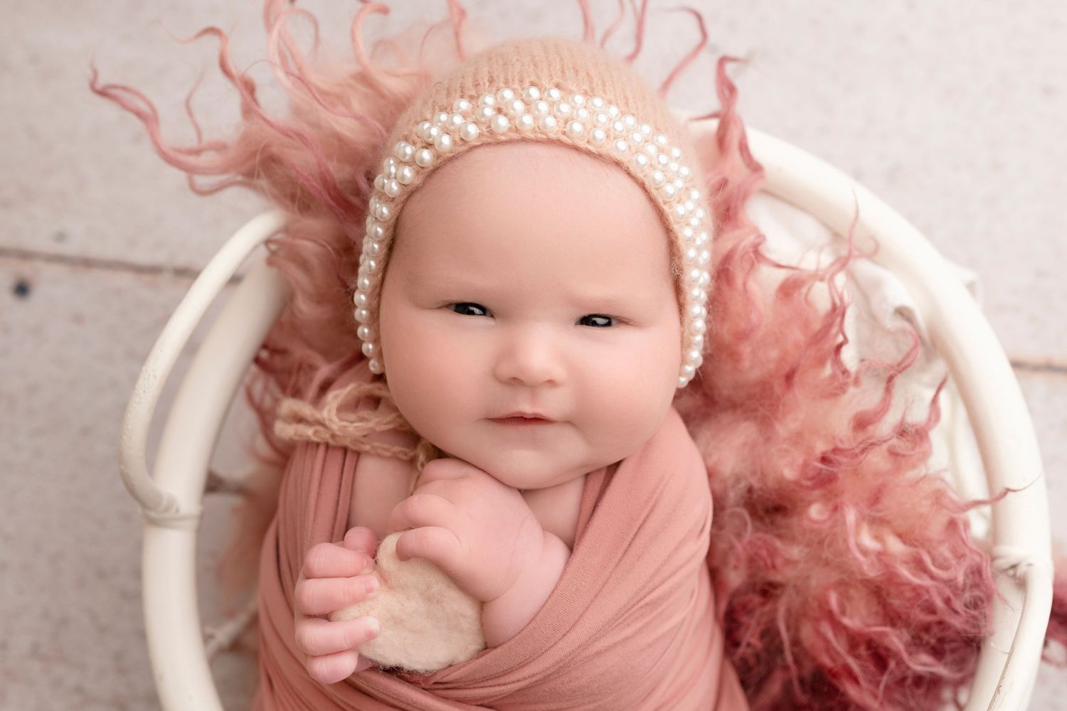 Newborn baby wrapped in pink swaddle with pearl bonnet, posed in white basket with fluffy pink fur.