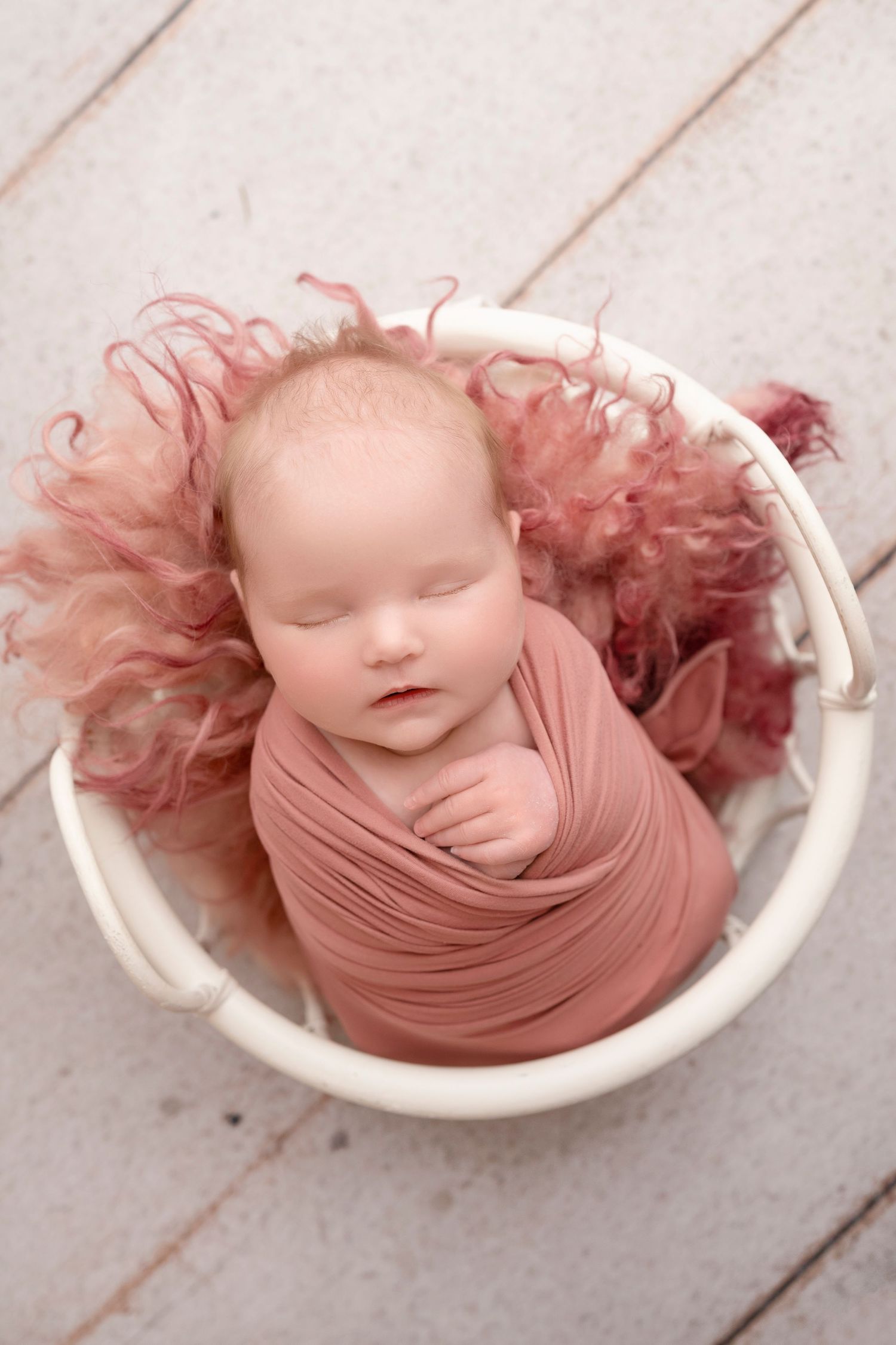 Sleeping newborn baby wrapped in dusty rose fabric nestled in a white bucket with pink fluffy blanket.
