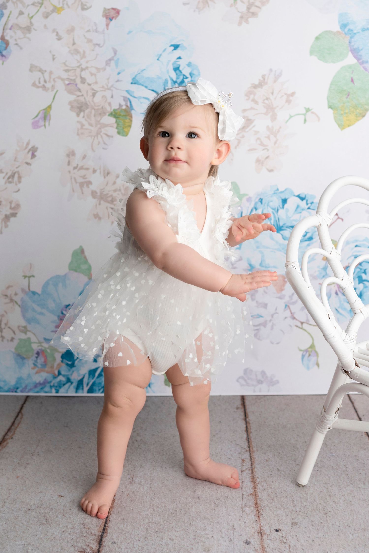 Baby girl in white tulle romper and blue bow headband stands barefoot before floral backdrop.