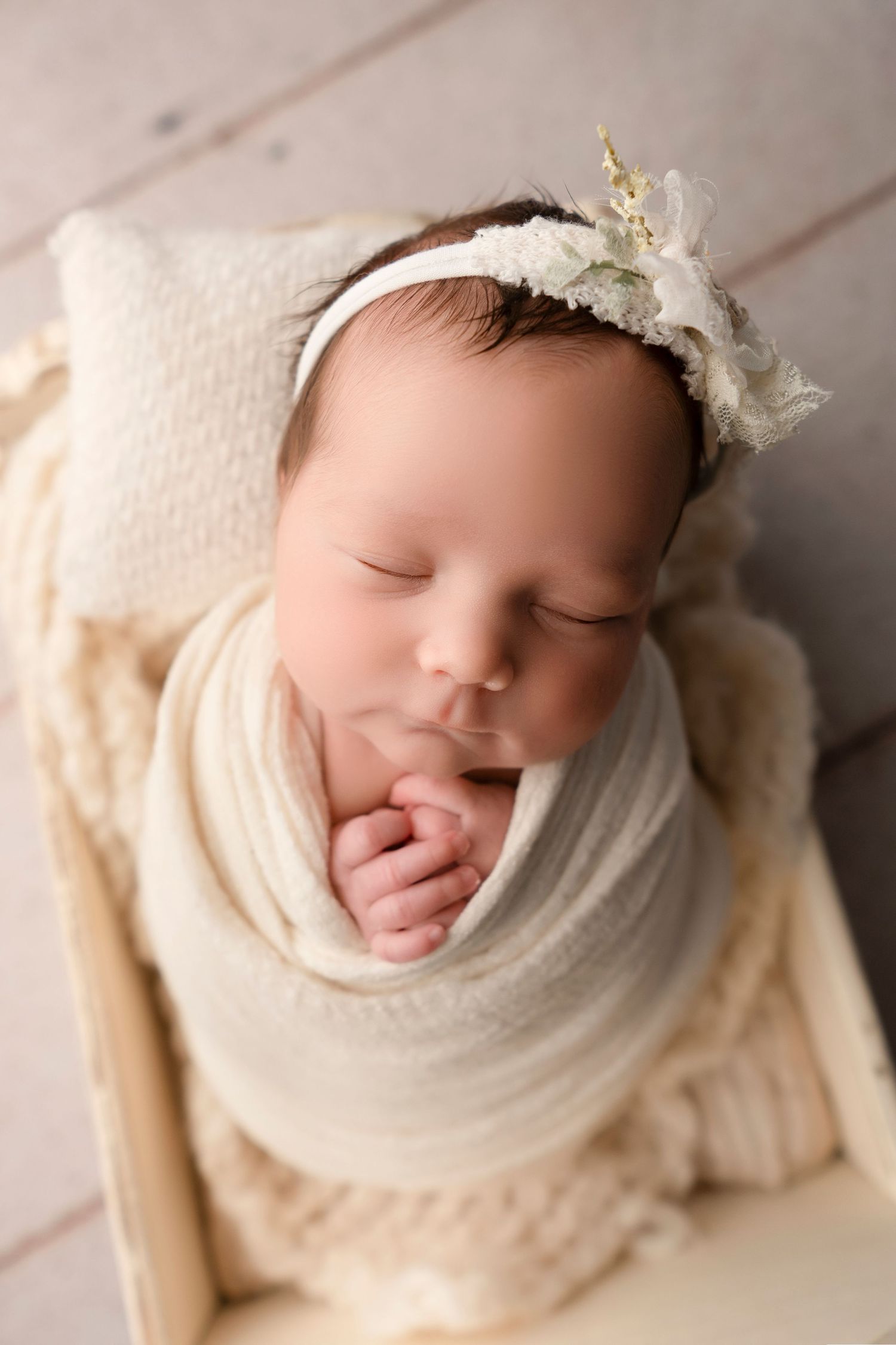 Sleeping newborn baby girl swaddled in cream wrap with floral headband resting in a wooden bed.