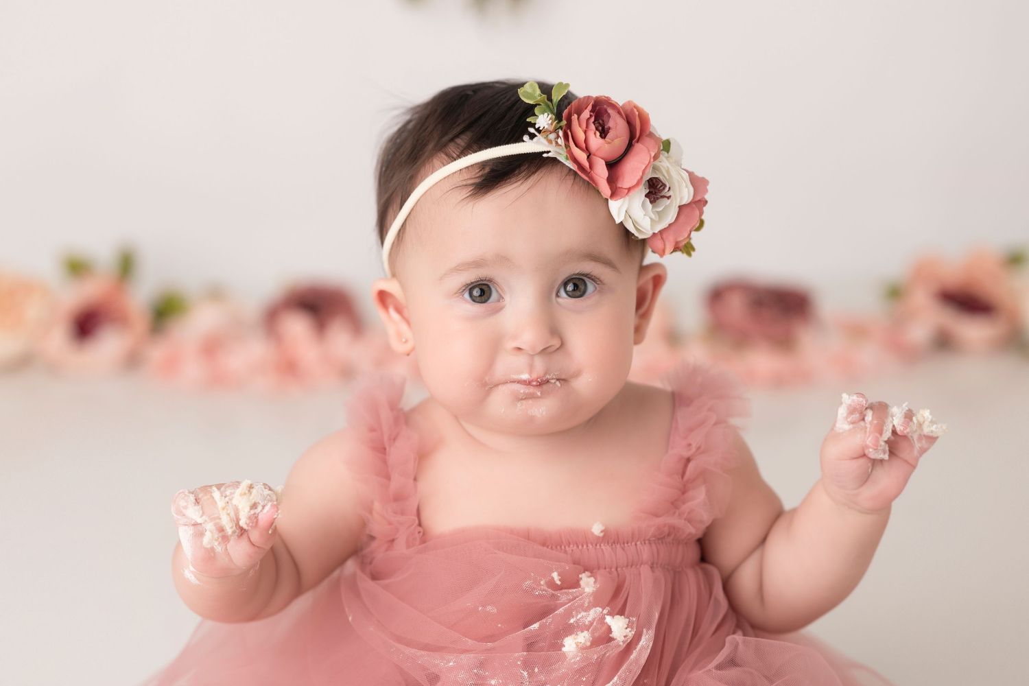 Baby in pink dress and floral headband with hands raised against floral backdrop.