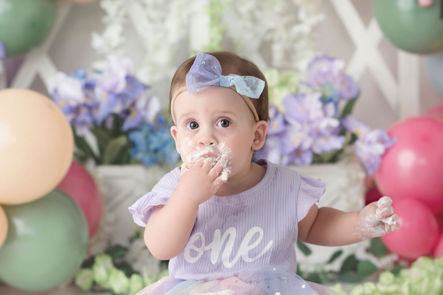 Baby girl in purple dress eating cake at her first birthday smash cake session with balloons and flowers.