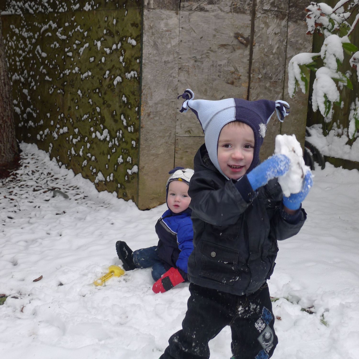 Two young children playing in the snow, one holding a snowball wearing a jester hat, another sitting in background in blue jacket.