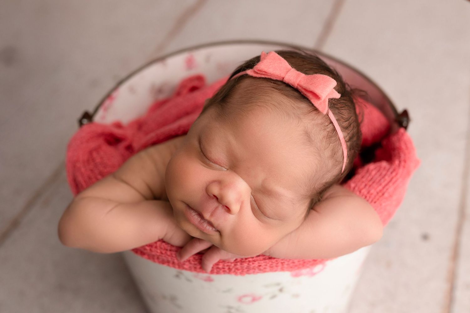 Peaceful newborn baby girl sleeping in a white floral bucket with pink knit wrap and bow headband.