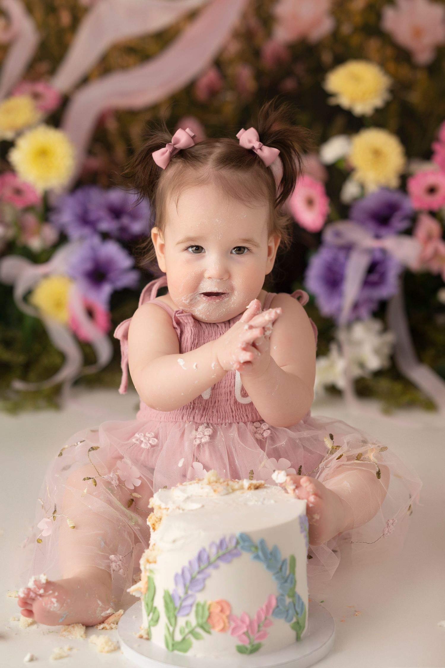 A baby girl in a pink tulle dress claps her hands at her first birthday cake smash session with floral backdrop.